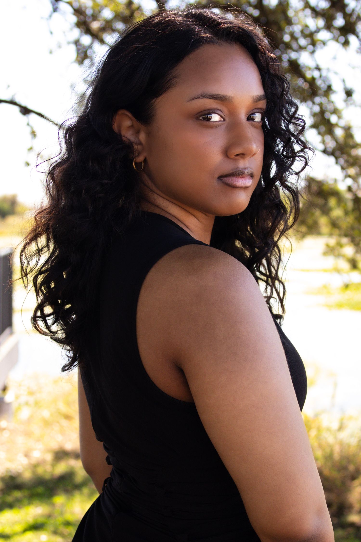 Young Black woman with curly hair in black sleeveless top looks over shoulder outdoors in natural light.