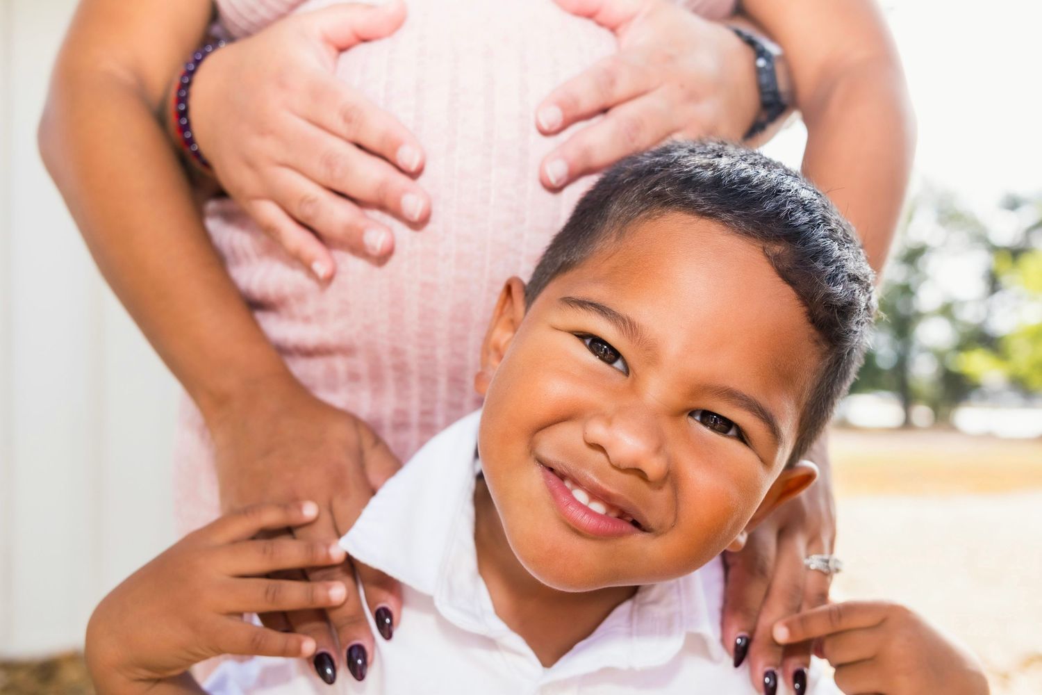Smiling young boy in front of pregnant mother's belly, family maternity photo outdoors.