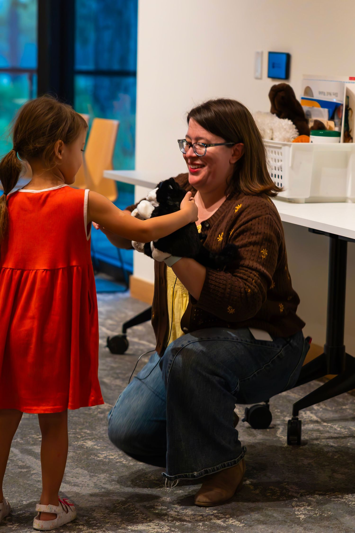 Woman crouching and smiling while young girl in red dress pets a black and white guinea pig indoors.