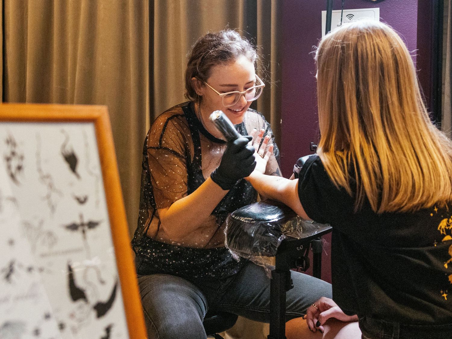 Tattoo artist in glasses applying ink on client in a studio with flash art displayed on the wall.