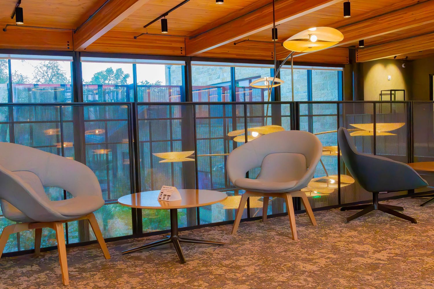 Modern lounge area with gray chairs, wooden tables, and floor-to-ceiling windows overlooking a courtyard.