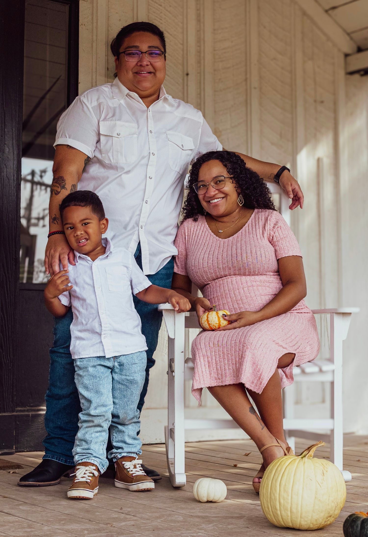 Family of three posing on a porch with pumpkins, pregnant woman in pink dress, man in white shirt, young boy in jeans.