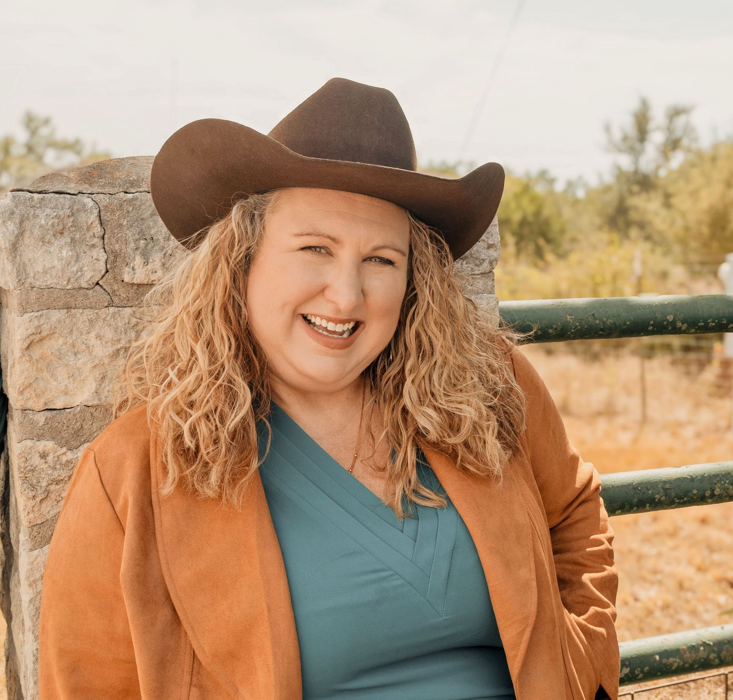 A cheerful woman in a brown cowboy hat and orange jacket smiles outdoors near a fence.