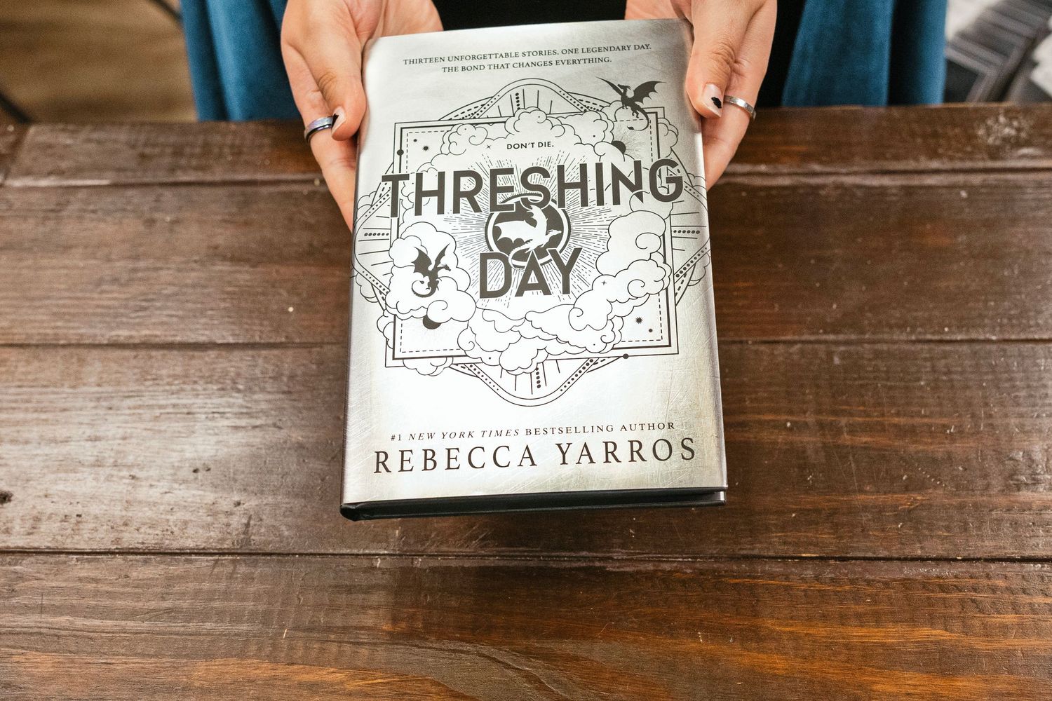 Hands holding a hardcover book titled Threshing Day by Rebecca Yarros on a wooden table.