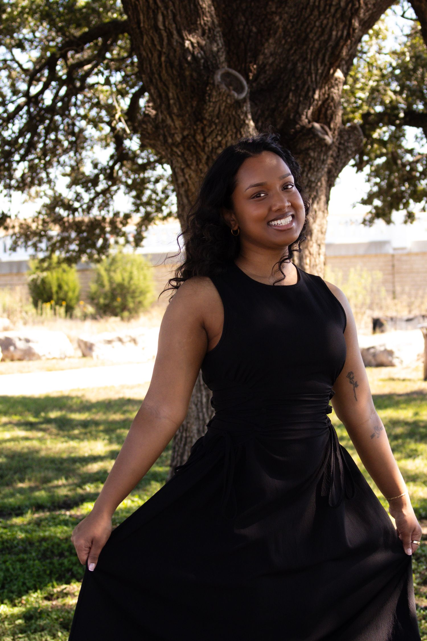 A smiling young Black woman in a black sleeveless maxi dress poses outdoors near a large tree on a sunny day.