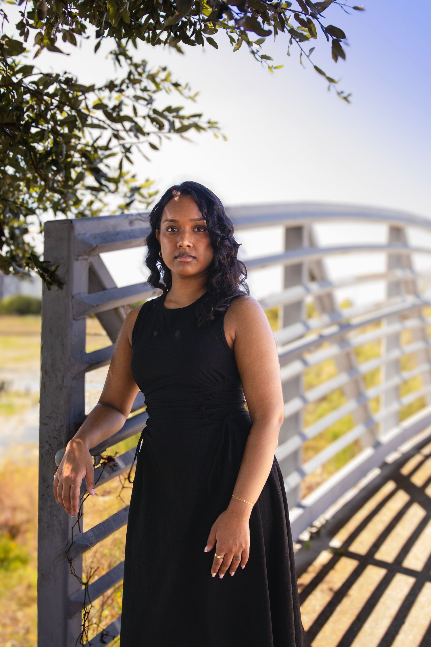Young woman in black dress poses by metal gate with tree branches overhead in outdoor portrait.