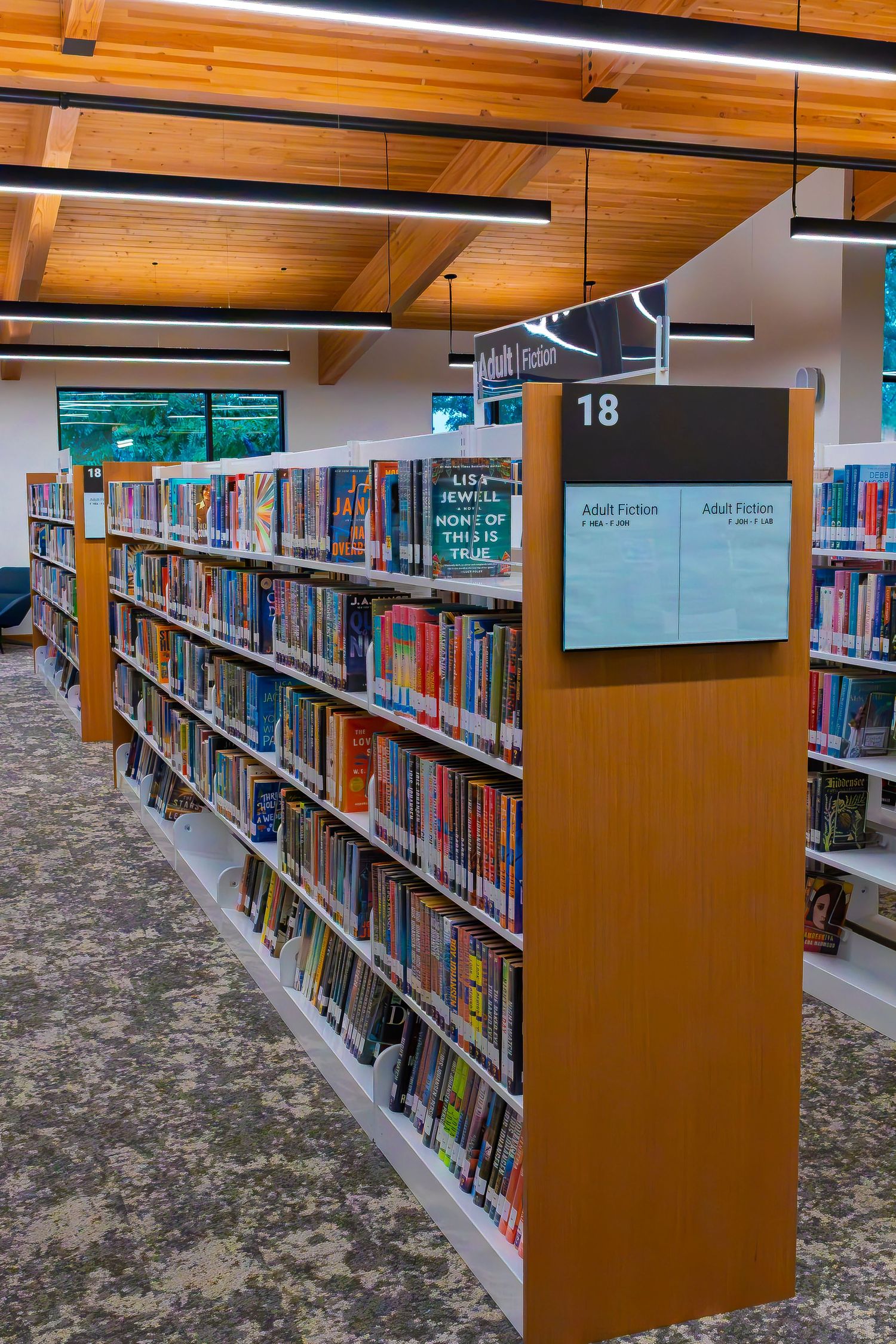 Library aisle with colorful books on shelves, section 18 Adult Fiction sign visible, modern ceiling with wooden beams.