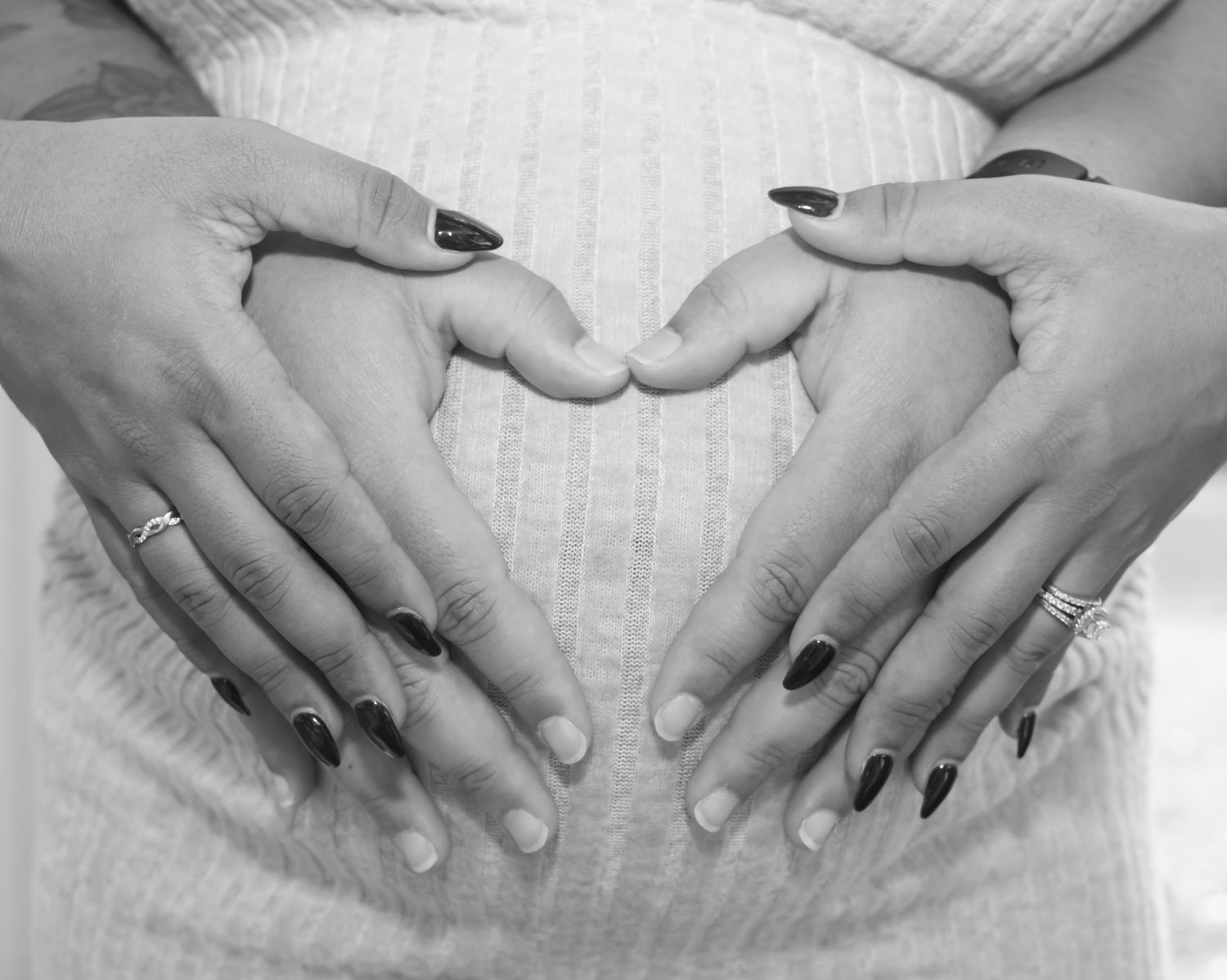 Black and white photo of two people forming a heart shape with their hands on a pregnant belly.