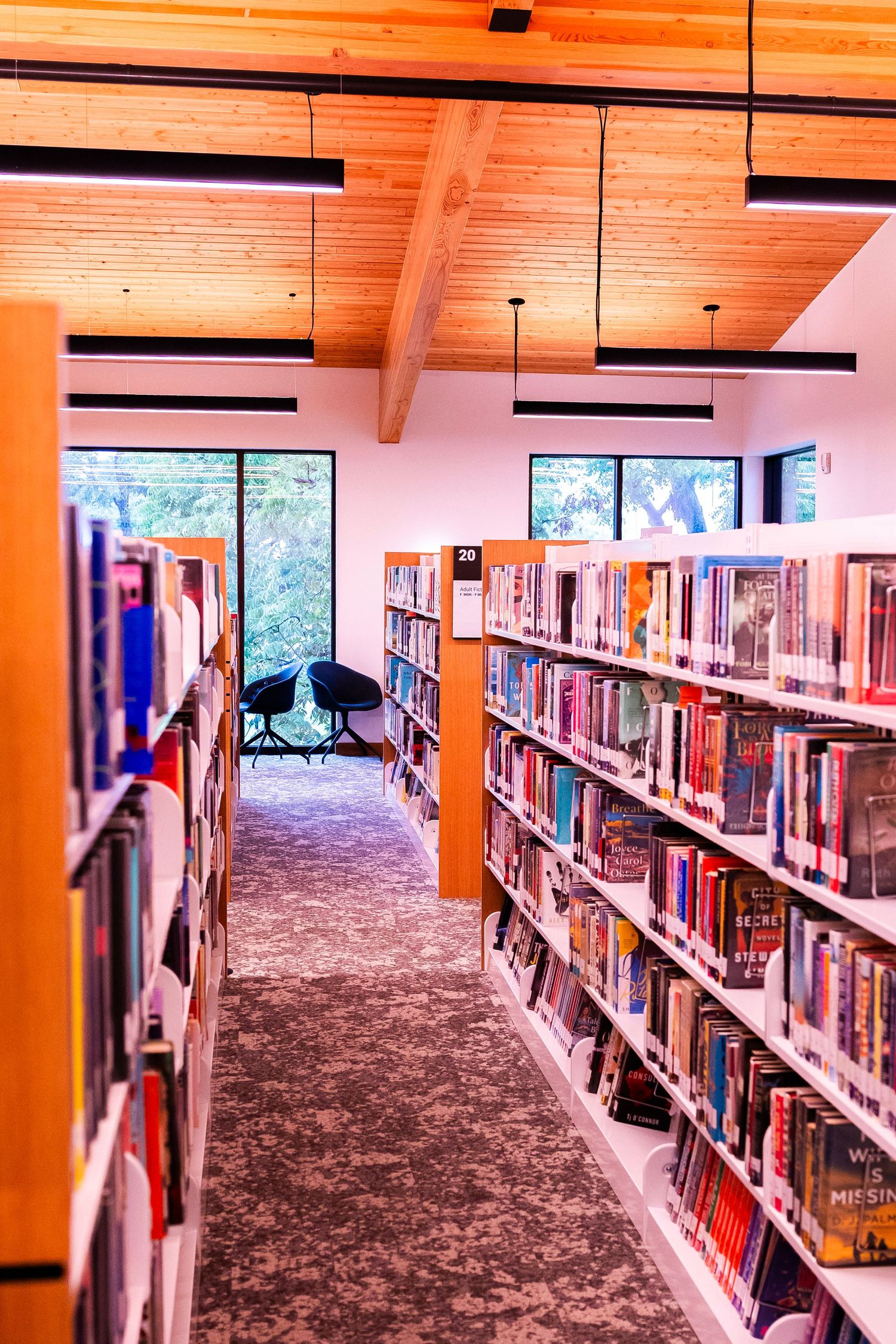 Modern library interior with wooden ceiling, pendant lights, and shelves filled with colorful books leading to cozy seating.