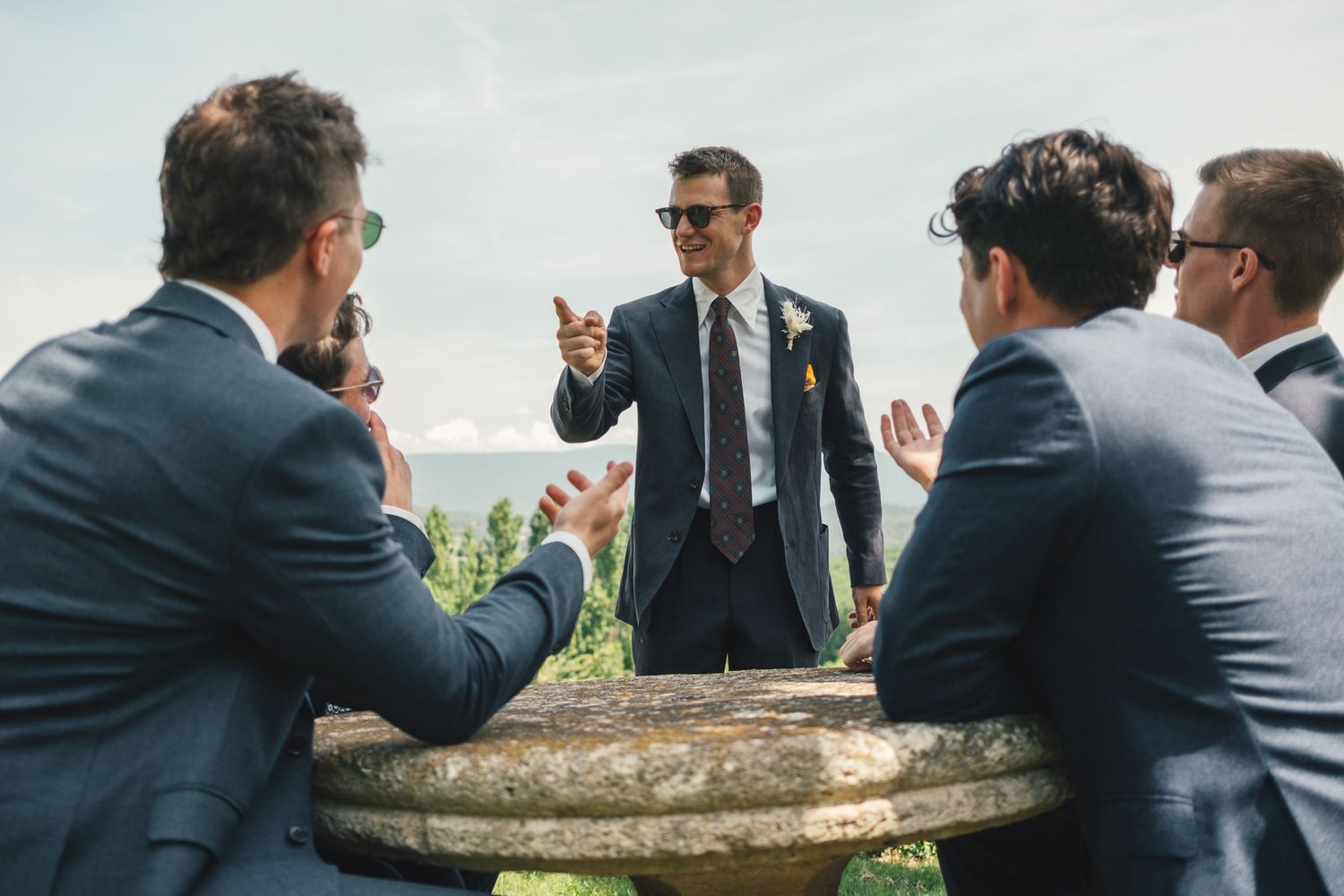 Groom in navy suit with boutonniere talks with groomsmen around stone table at outdoor wedding.