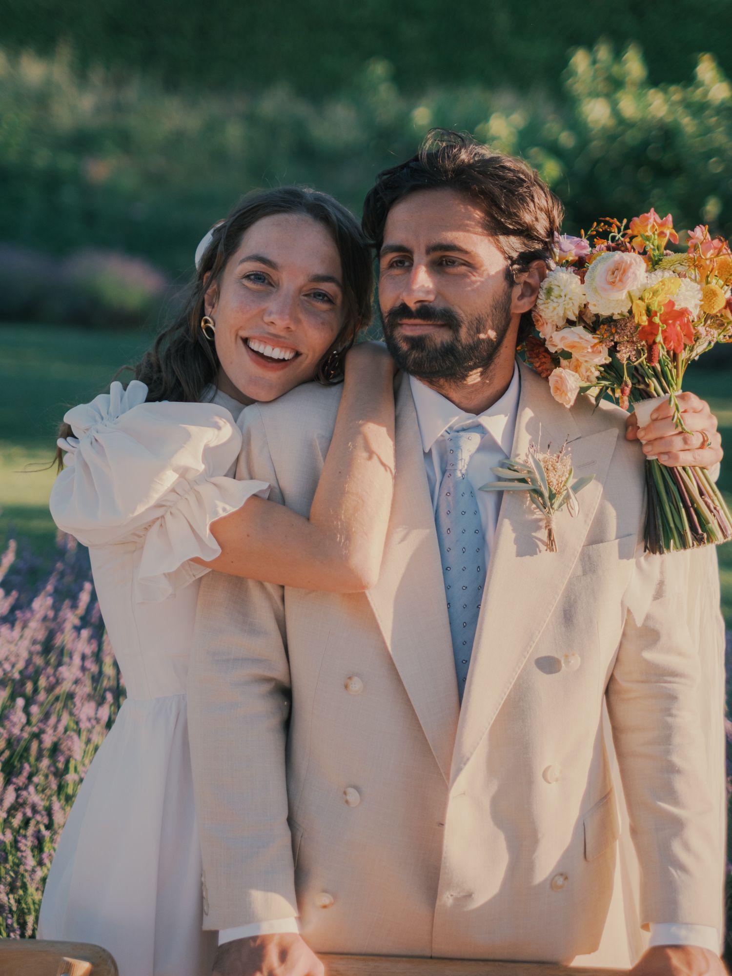 Bride and groom in beige suit pose together holding a colorful bouquet in a lush garden setting.