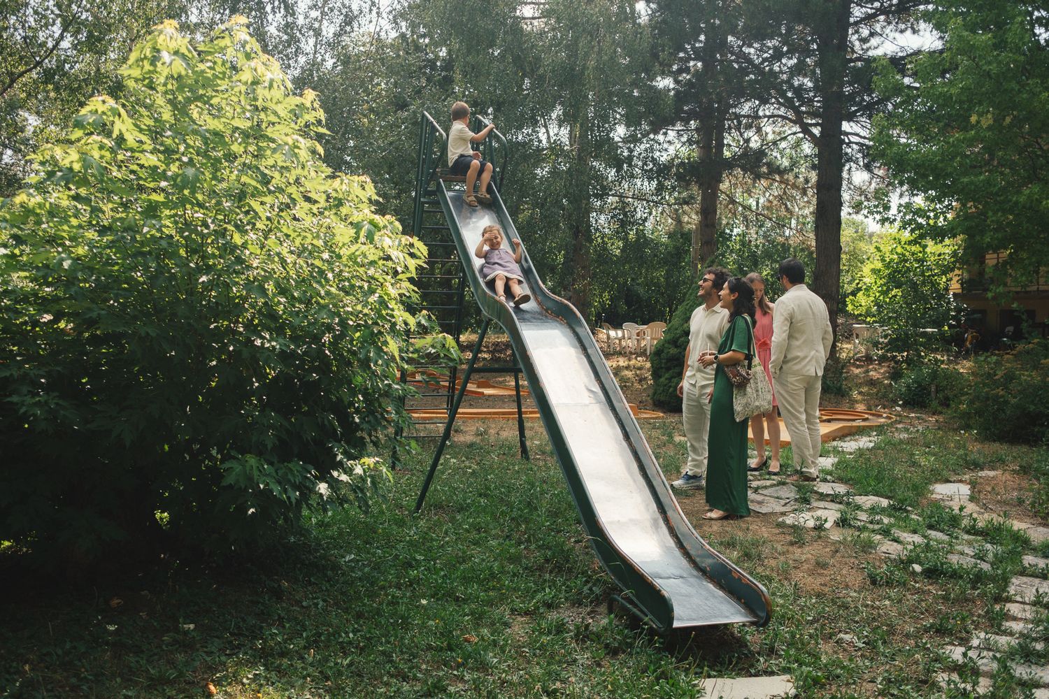 Children playing on a backyard metal slide surrounded by lush trees while adults watch nearby.