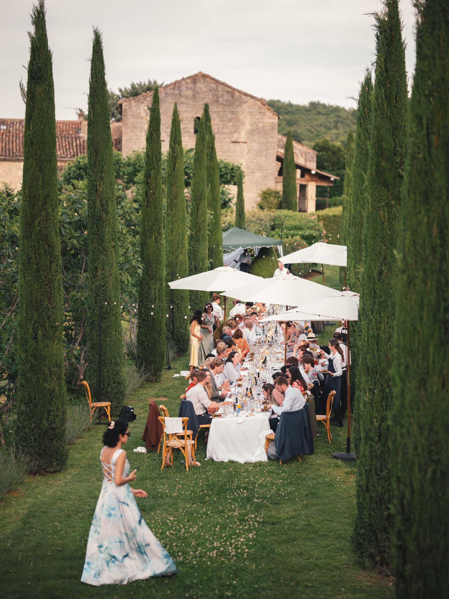 Elegant outdoor wedding reception with long dining table, white umbrellas, and tall cypress trees at a Tuscan villa.