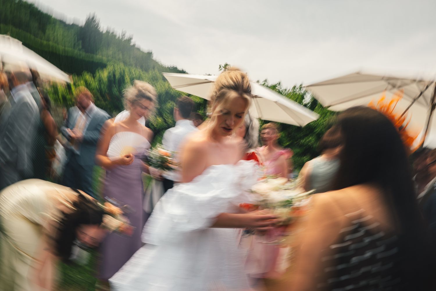 Bride in white gown walks through wedding guests outdoors, surrounded by colorful bouquets on a cloudy day.