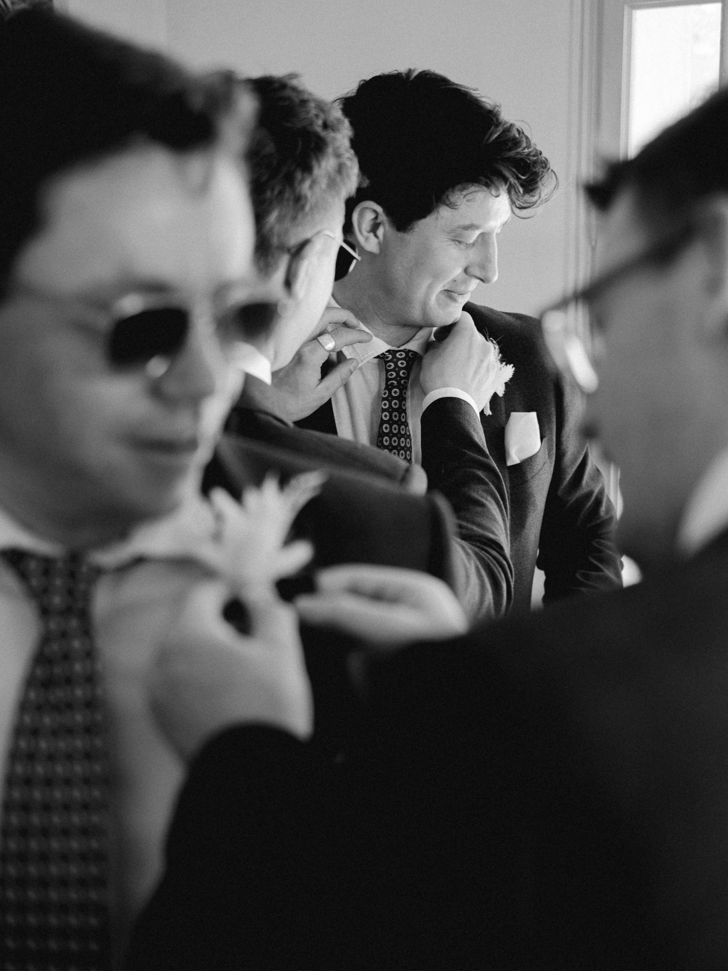 Black and white photo of groomsmen in suits adjusting ties and boutonnieres before a wedding ceremony.