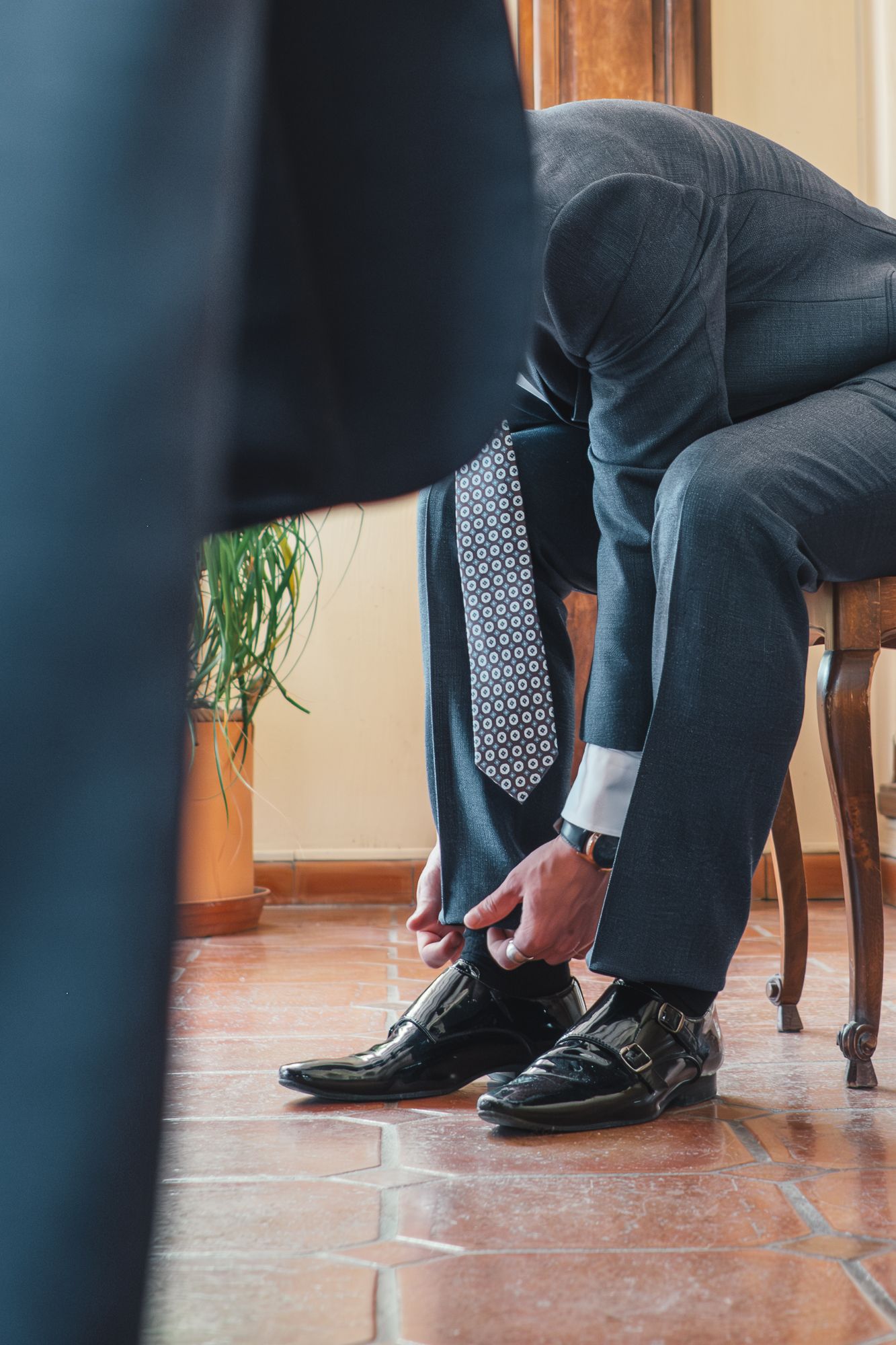 Man in suit and patterned tie fastening black monk strap shoes while seated on wooden chair.