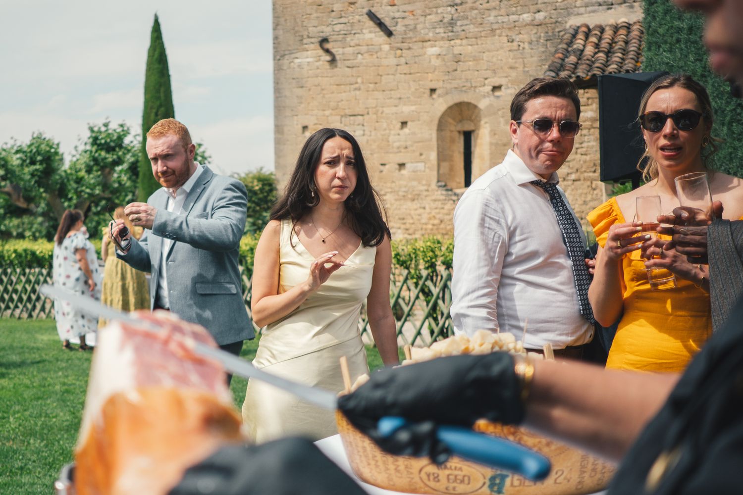 Wedding guests in formal attire socializing outdoors near a stone building on a sunny day.