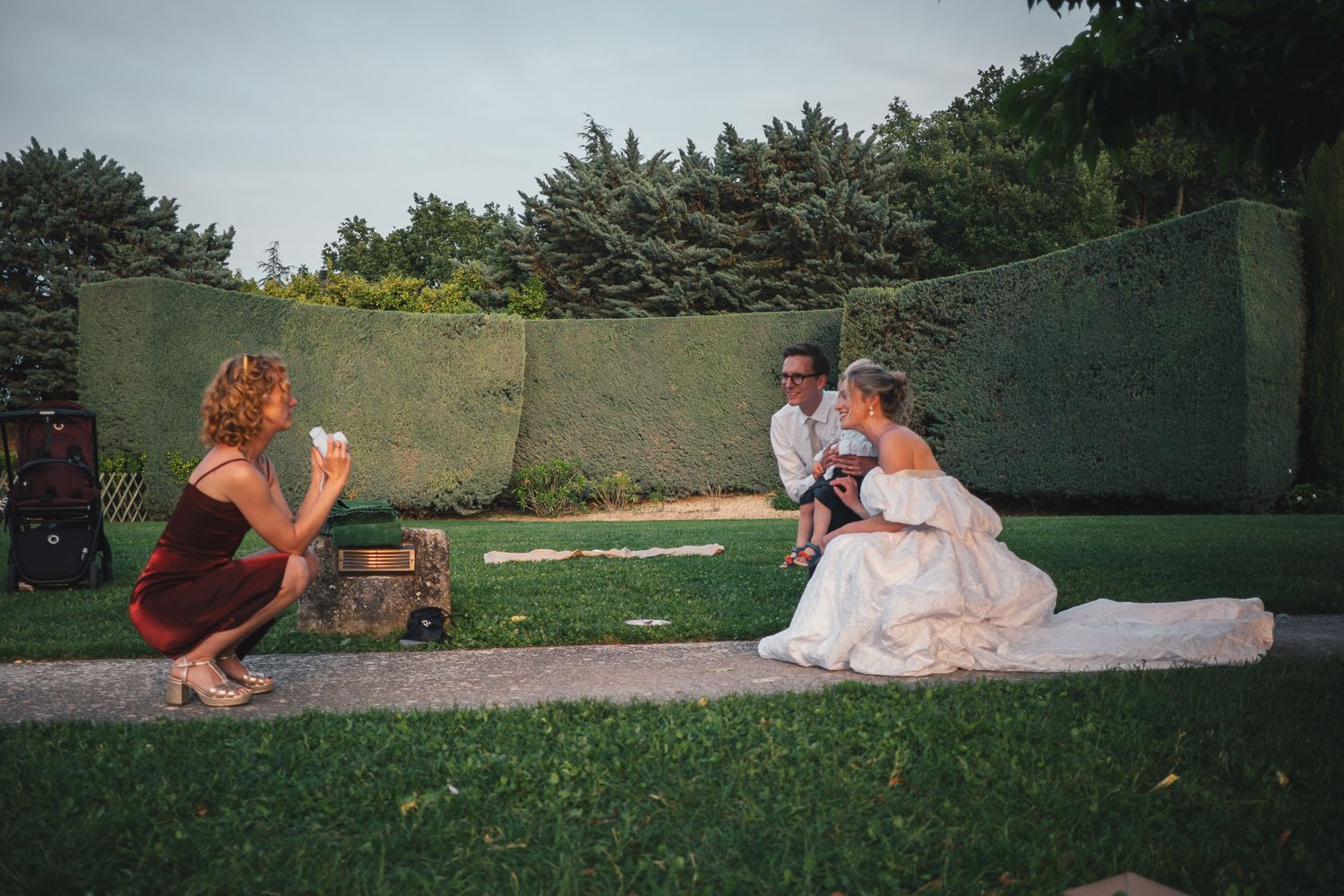 Woman photographs bride and groom sitting on grass in elegant garden during outdoor wedding photography session.
