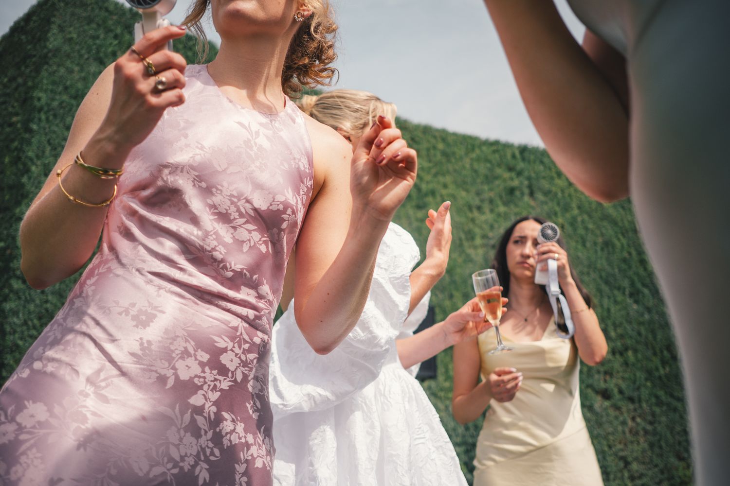 Women in elegant dresses blowing bubbles outdoors at a wedding celebration near a green hedge wall.