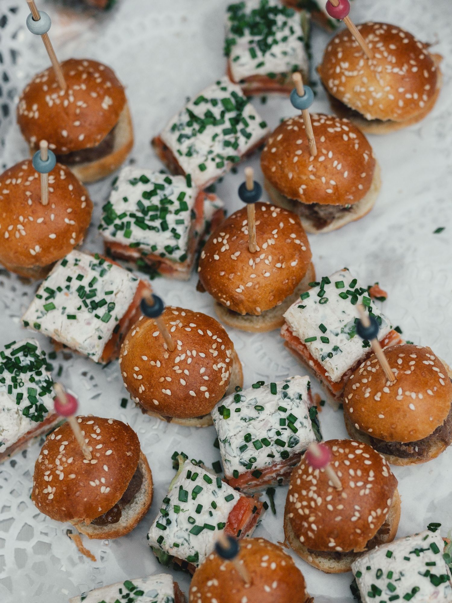 Assorted mini sliders and cream cheese appetizers on sesame buns, garnished with chives, served on a decorative platter.