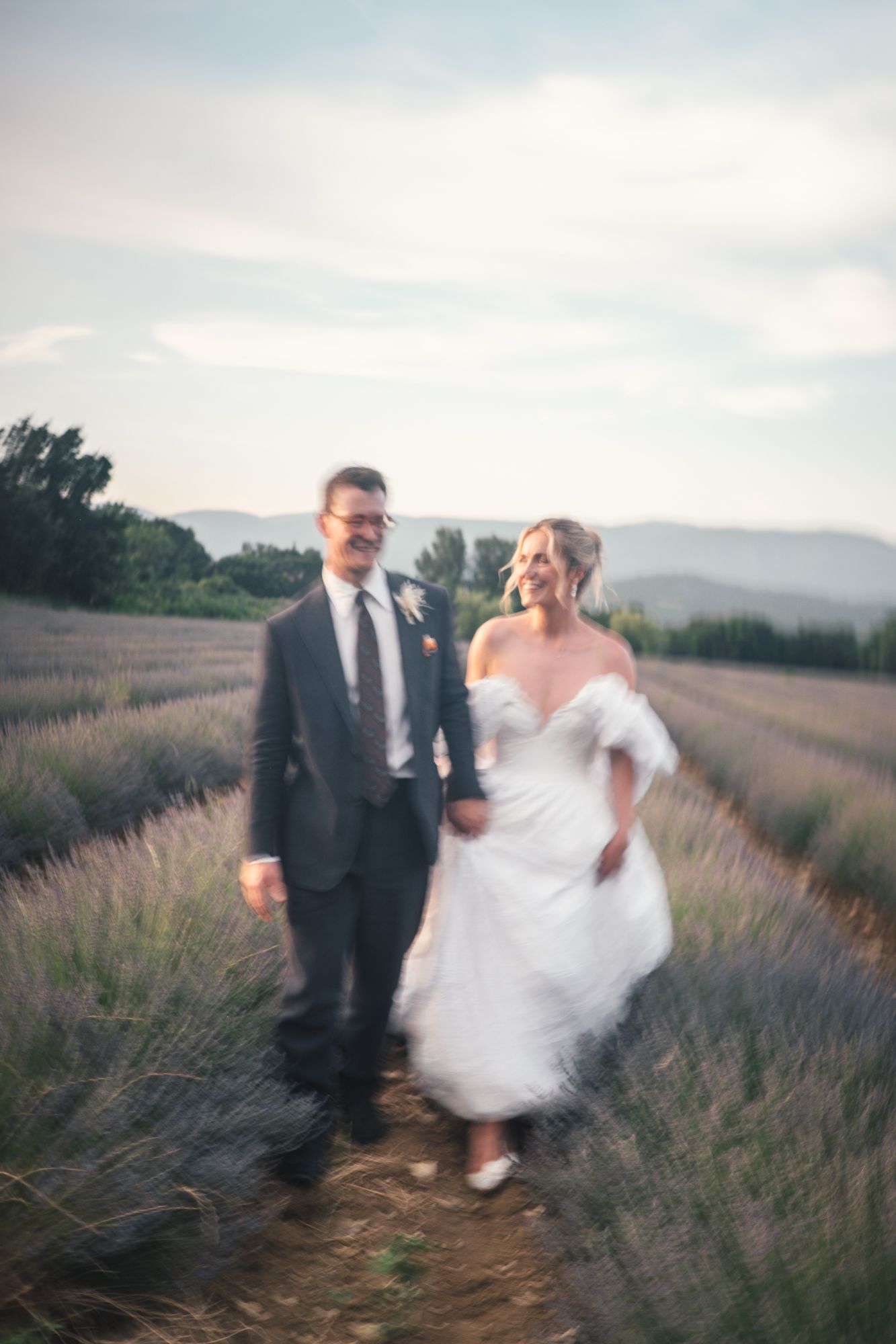 Bride and groom walk hand in hand through blooming lavender field at sunset in romantic motion blur photo.