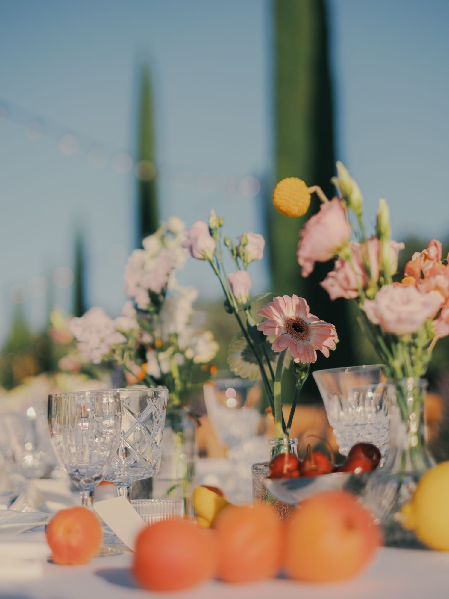 Elegant outdoor wedding table with crystal glasses, pink flowers, and fresh peaches under string lights.
