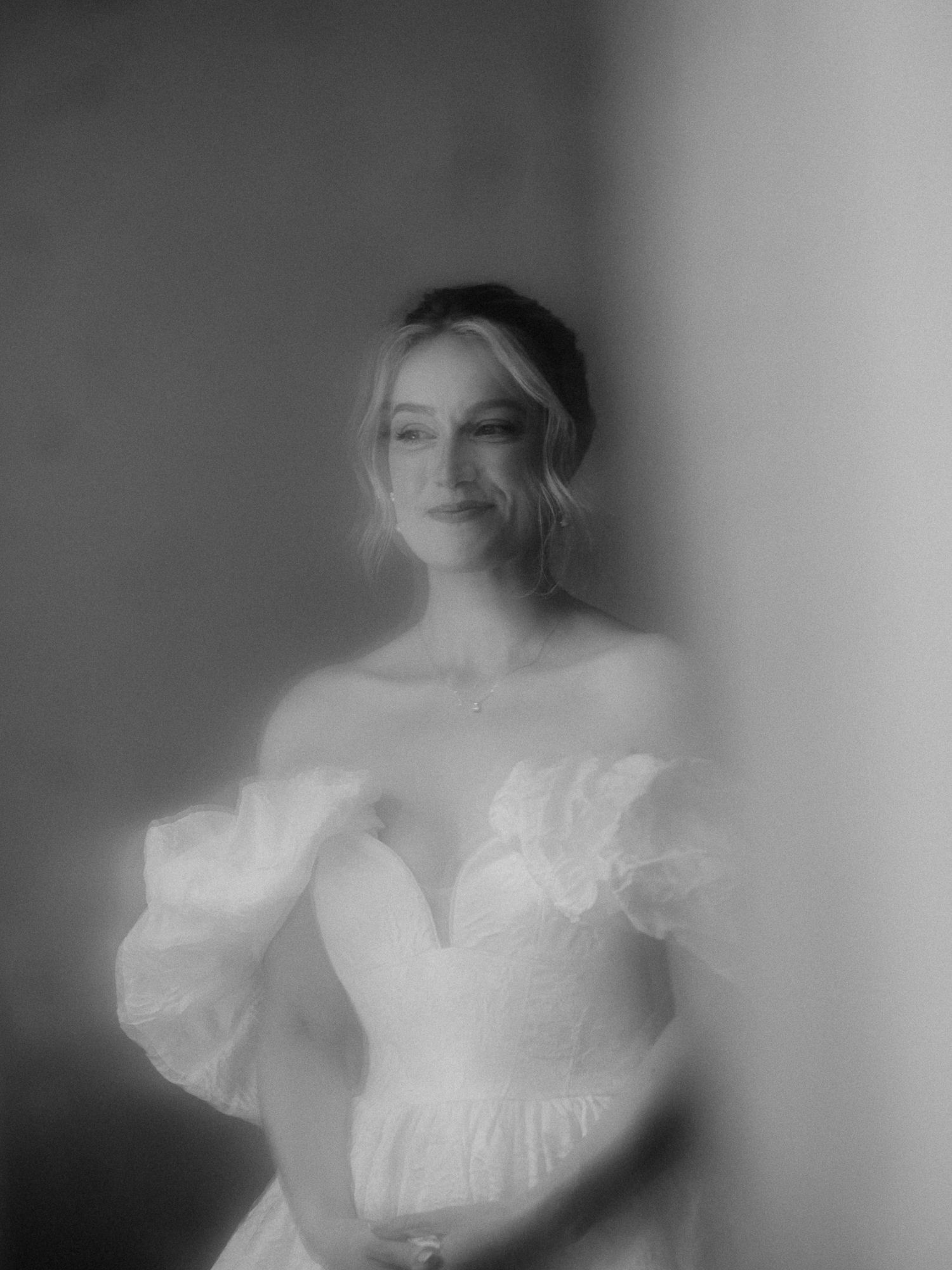 Dreamy black and white portrait of a bride in an off-shoulder ruffled wedding gown, softly lit against a neutral background.