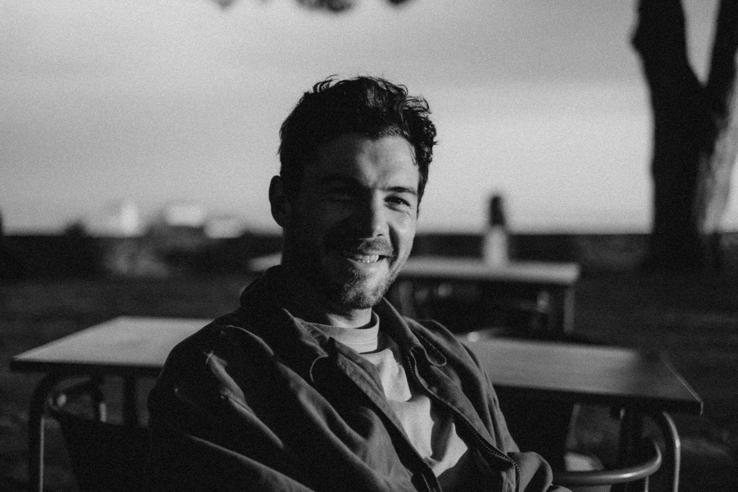 Smiling young man with curly hair sitting outdoors at a table in a black and white portrait photo.