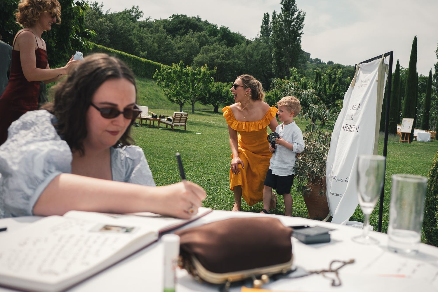 Guests sign wedding guestbook at outdoor table in lush green garden setting during summer wedding reception.