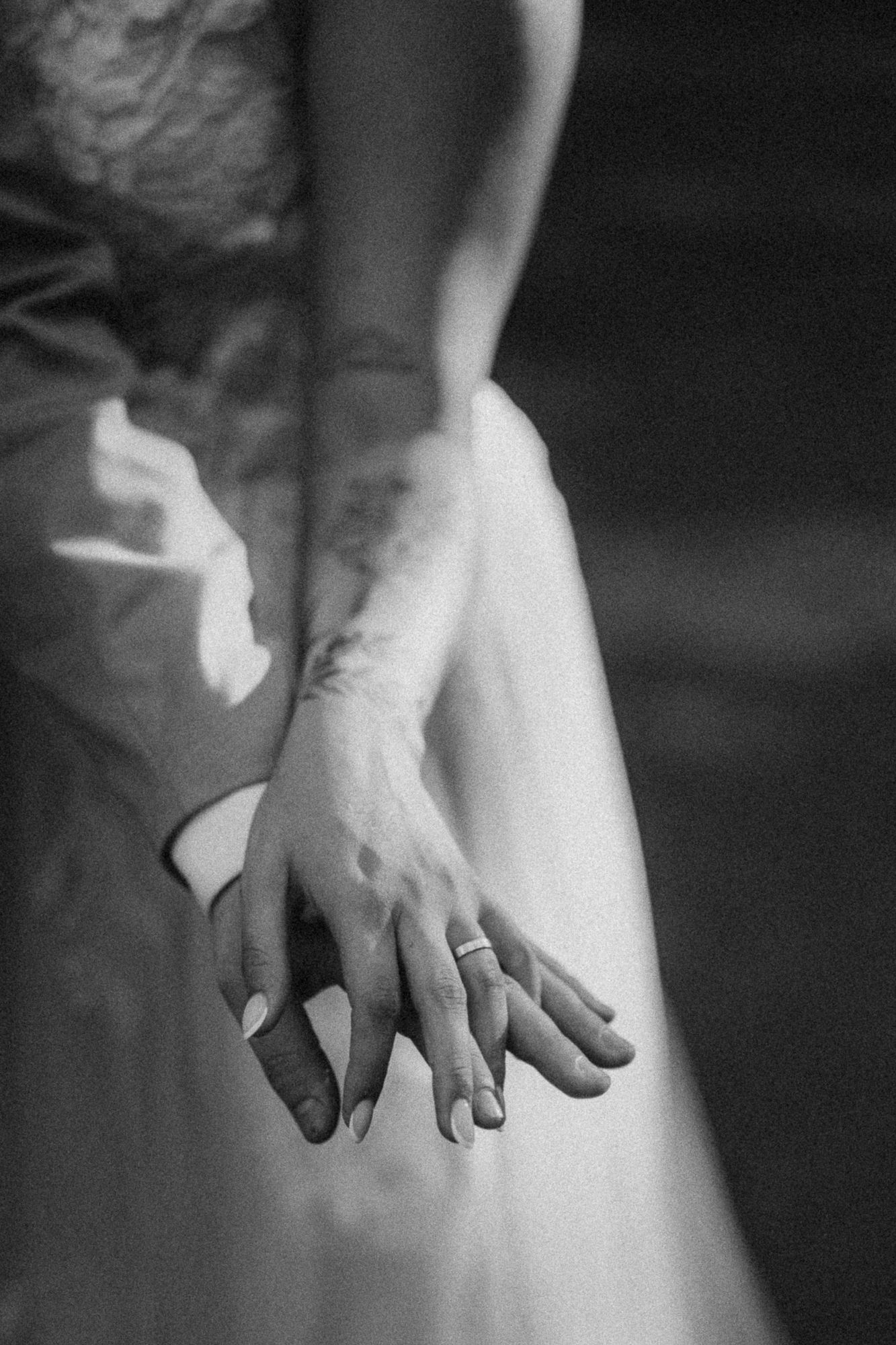 Black and white close-up of bride and groom holding hands showing wedding rings against white gown.