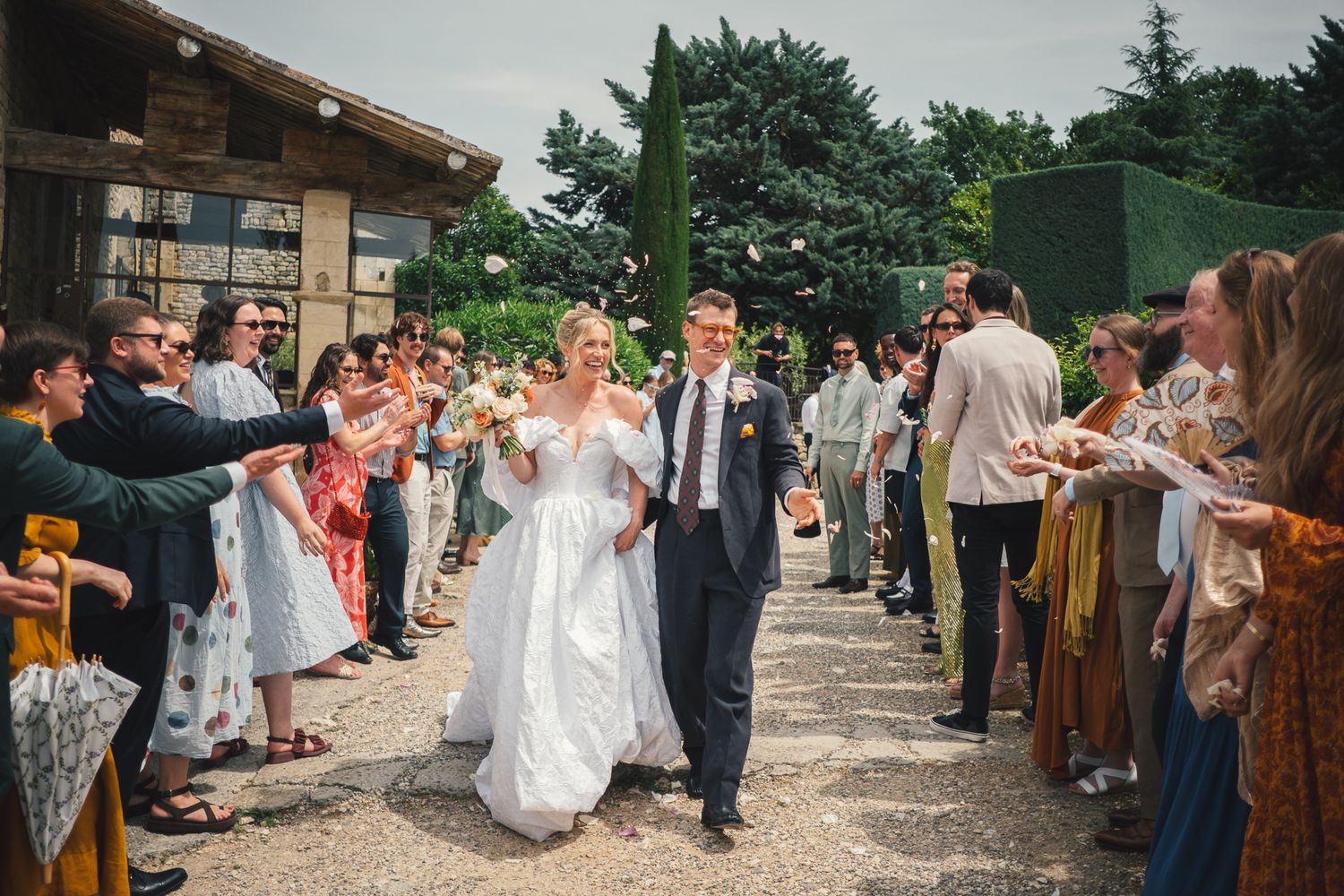 Bride and groom walk through confetti tunnel of guests at outdoor wedding venue on sunny day.