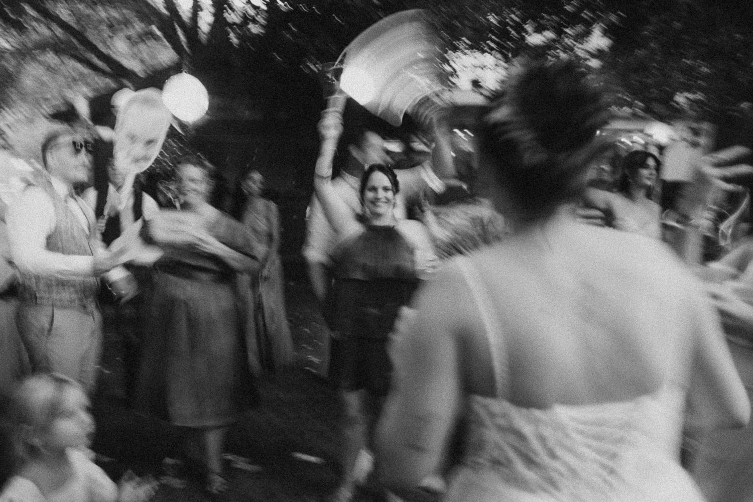 Black and white wedding celebration photo with guests cheering and bride in white dress at outdoor reception.