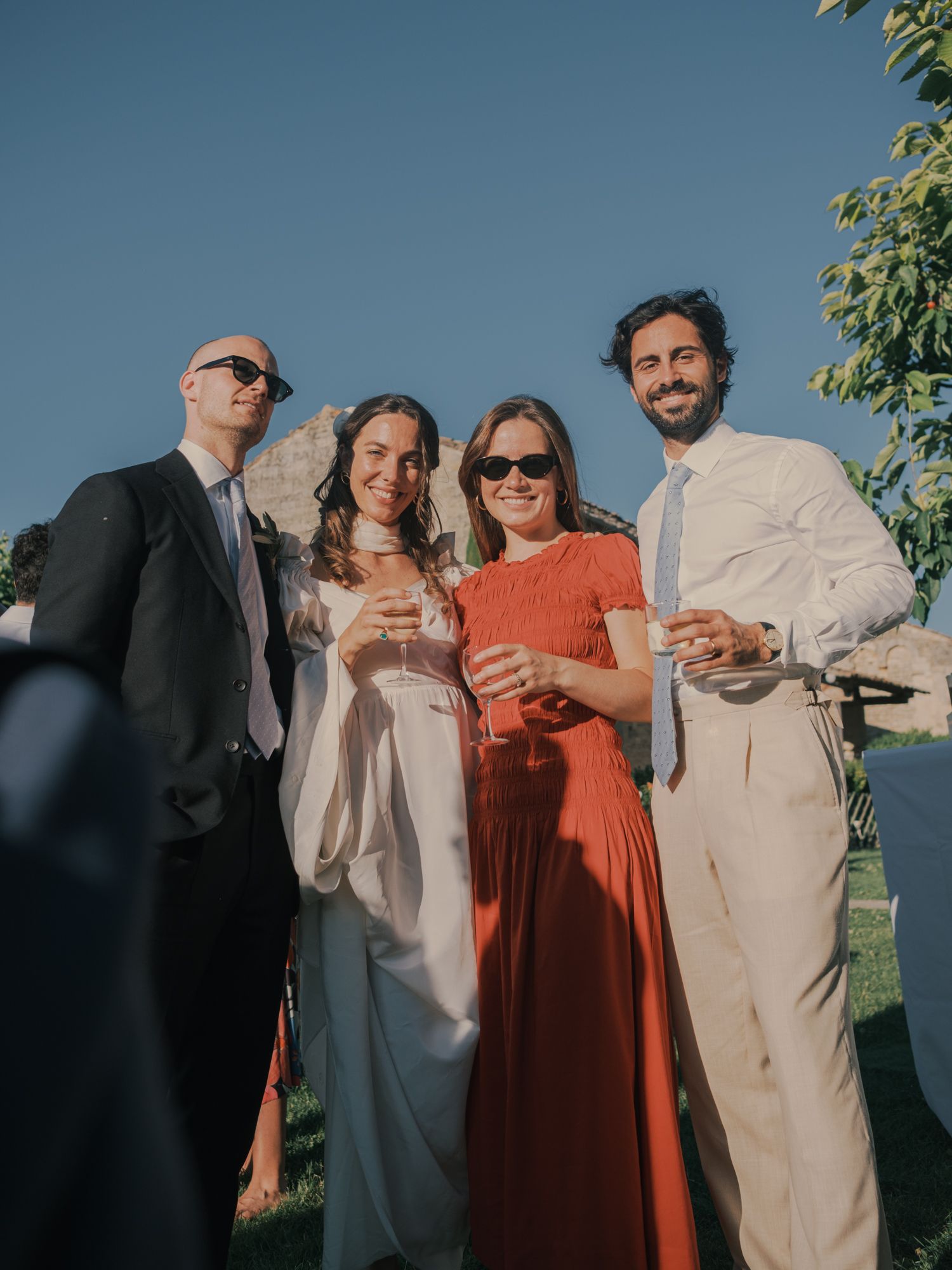 Four elegantly dressed wedding guests smiling and holding champagne glasses outdoors on a sunny day.