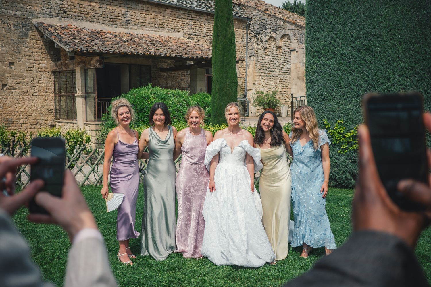 Bride poses with bridesmaids in elegant gowns outside a rustic stone villa, as guests photograph the group.