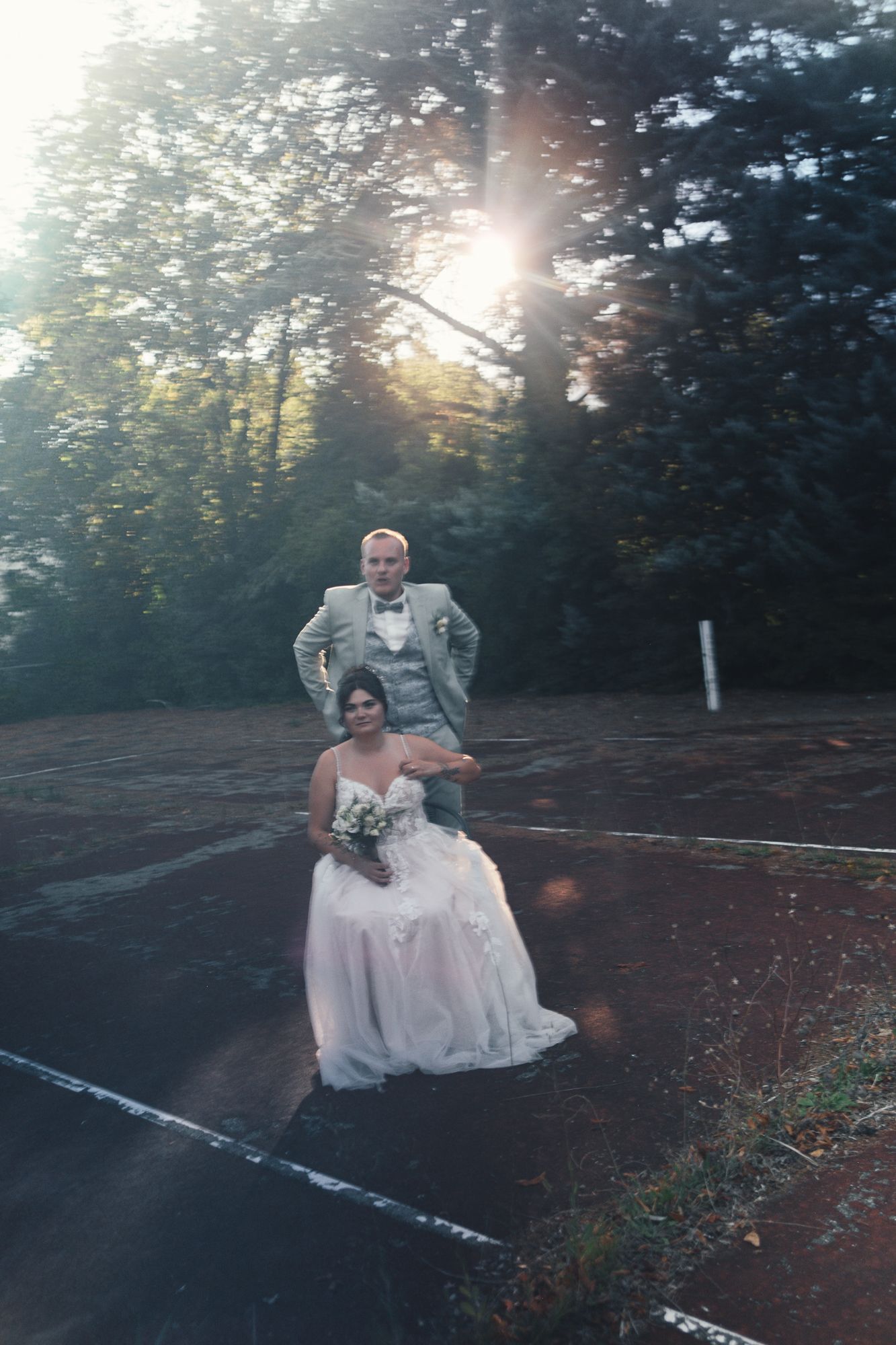 Bride in white gown seated outdoors with groom in gray suit, sunlight filtering through trees at golden hour.