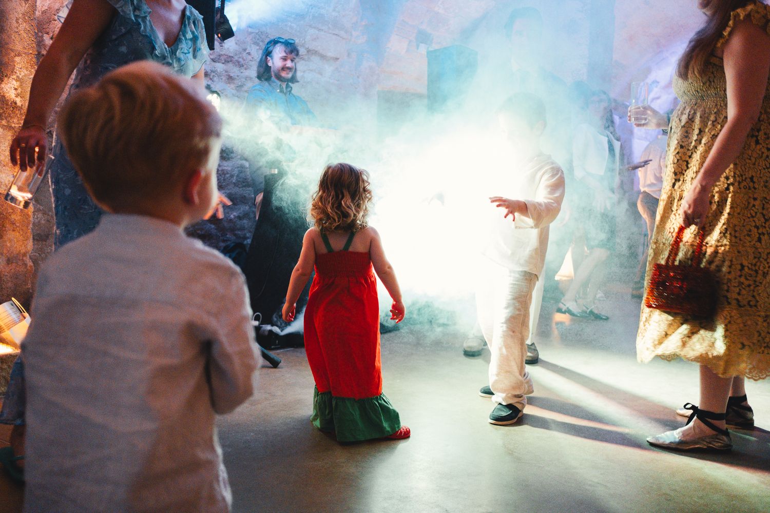 Children dancing in colorful outfits at a festive party with smoke effects and guests in the background.