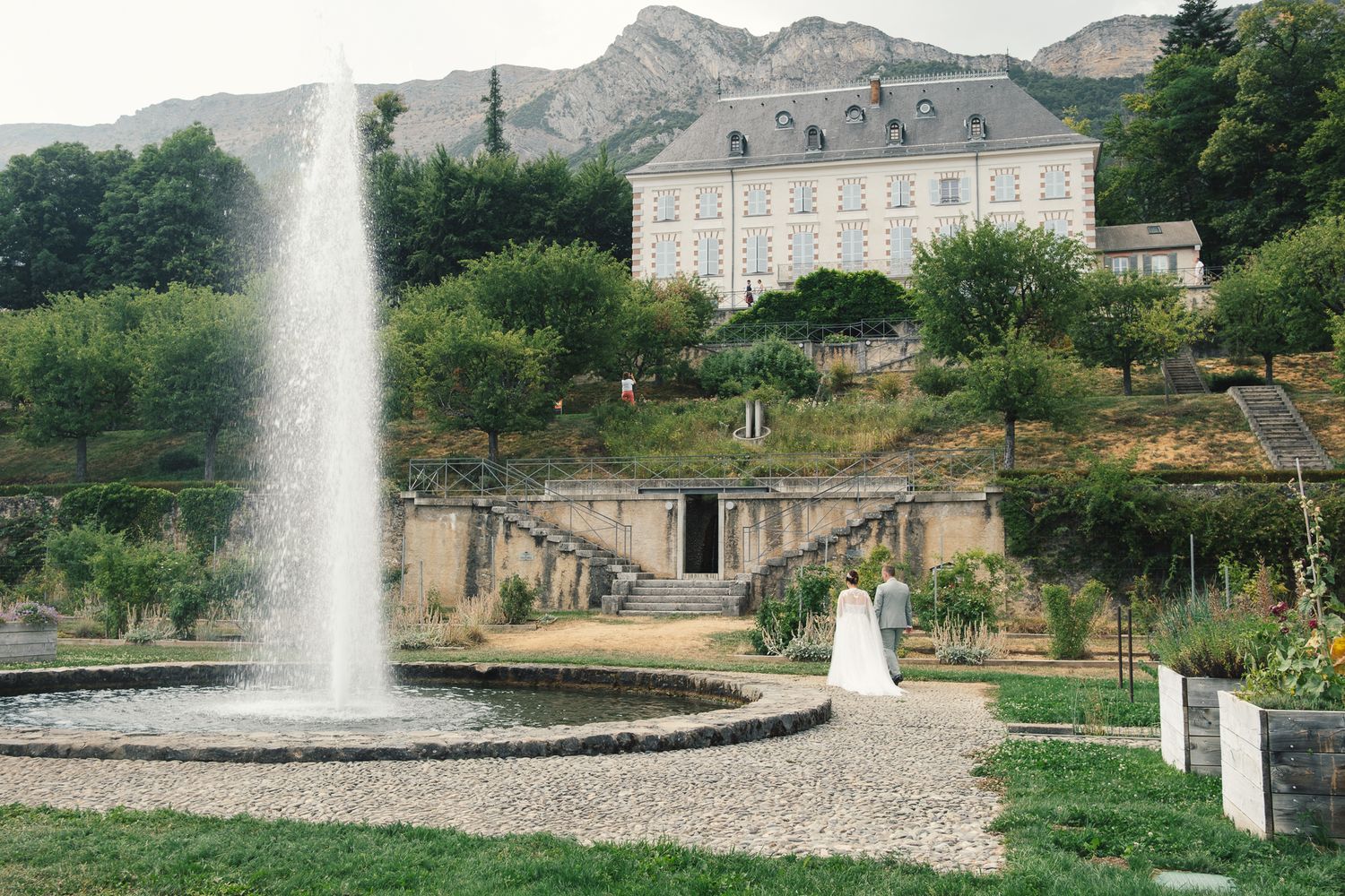 Elegant wedding couple near a grand fountain with a historic chateau and mountain backdrop.