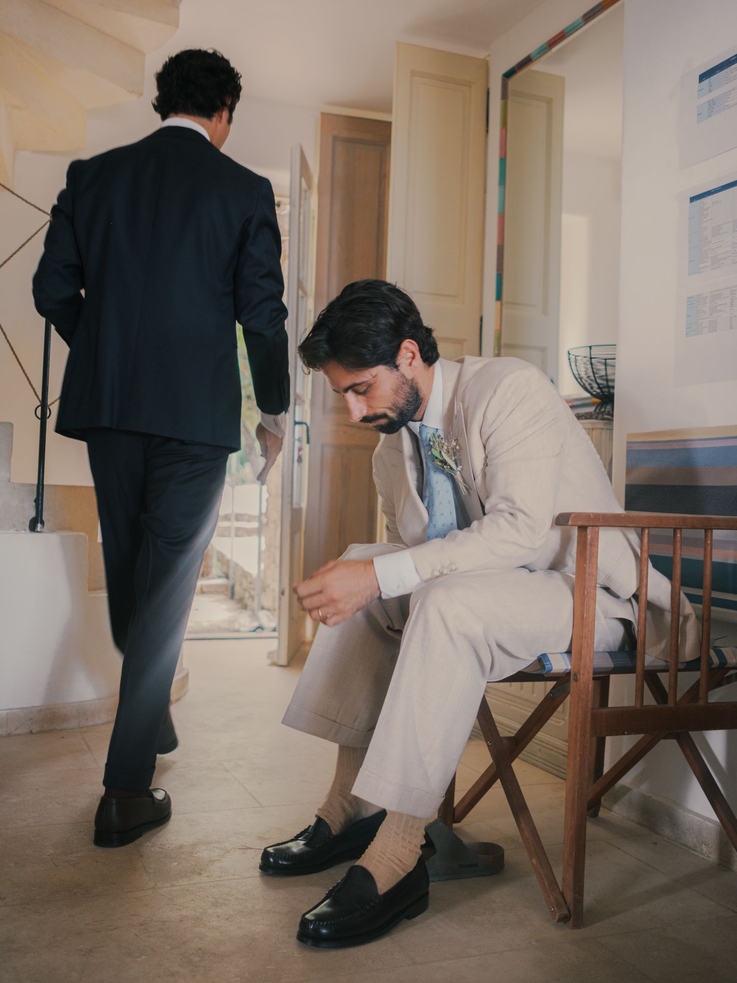 Two men in formal suits, one seated adjusting his loafer shoes, another walking away in a dark navy suit.