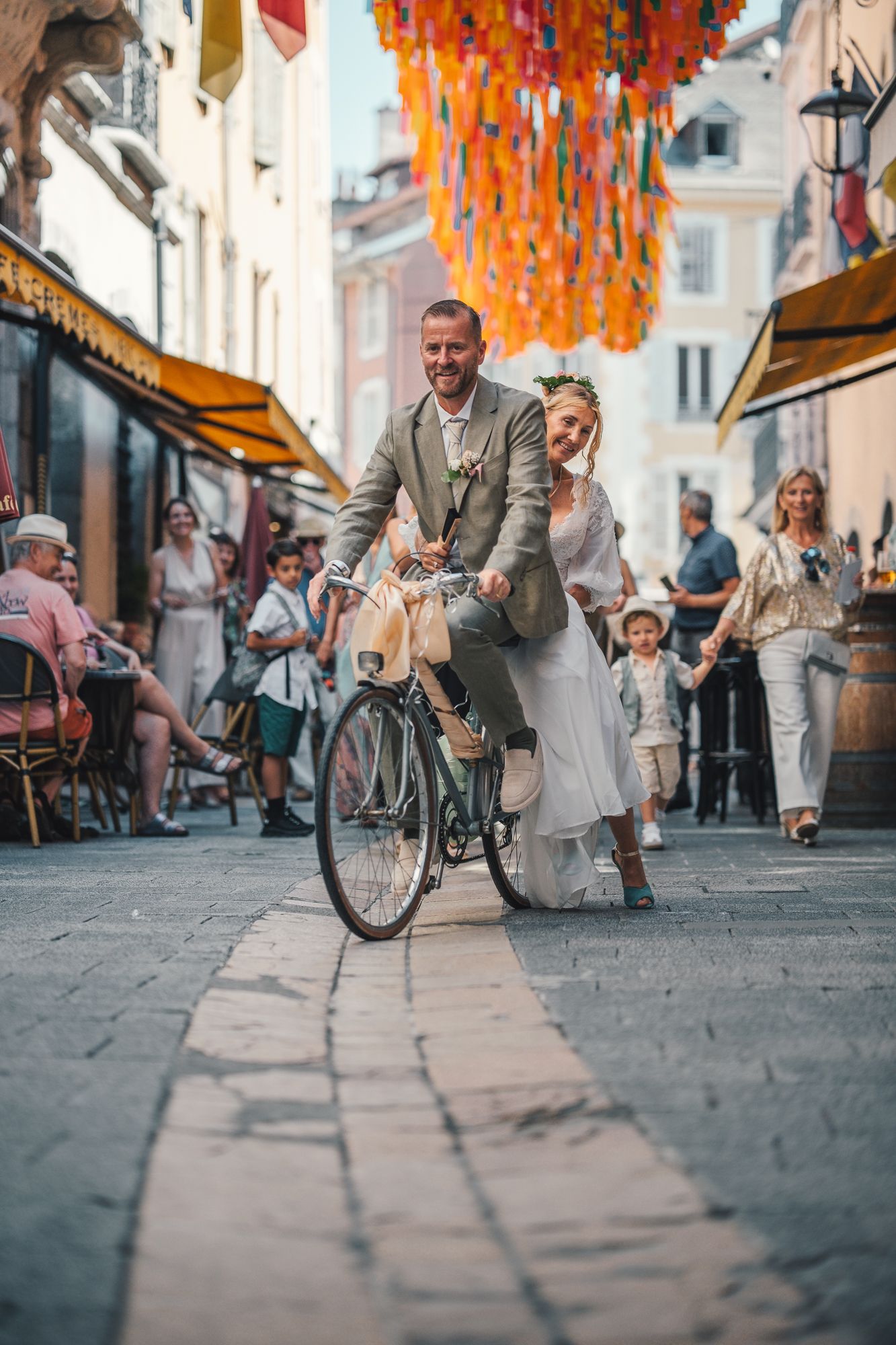 Bride and groom riding a vintage bicycle through a festive cobblestone street lined with cafes and colorful decorations.