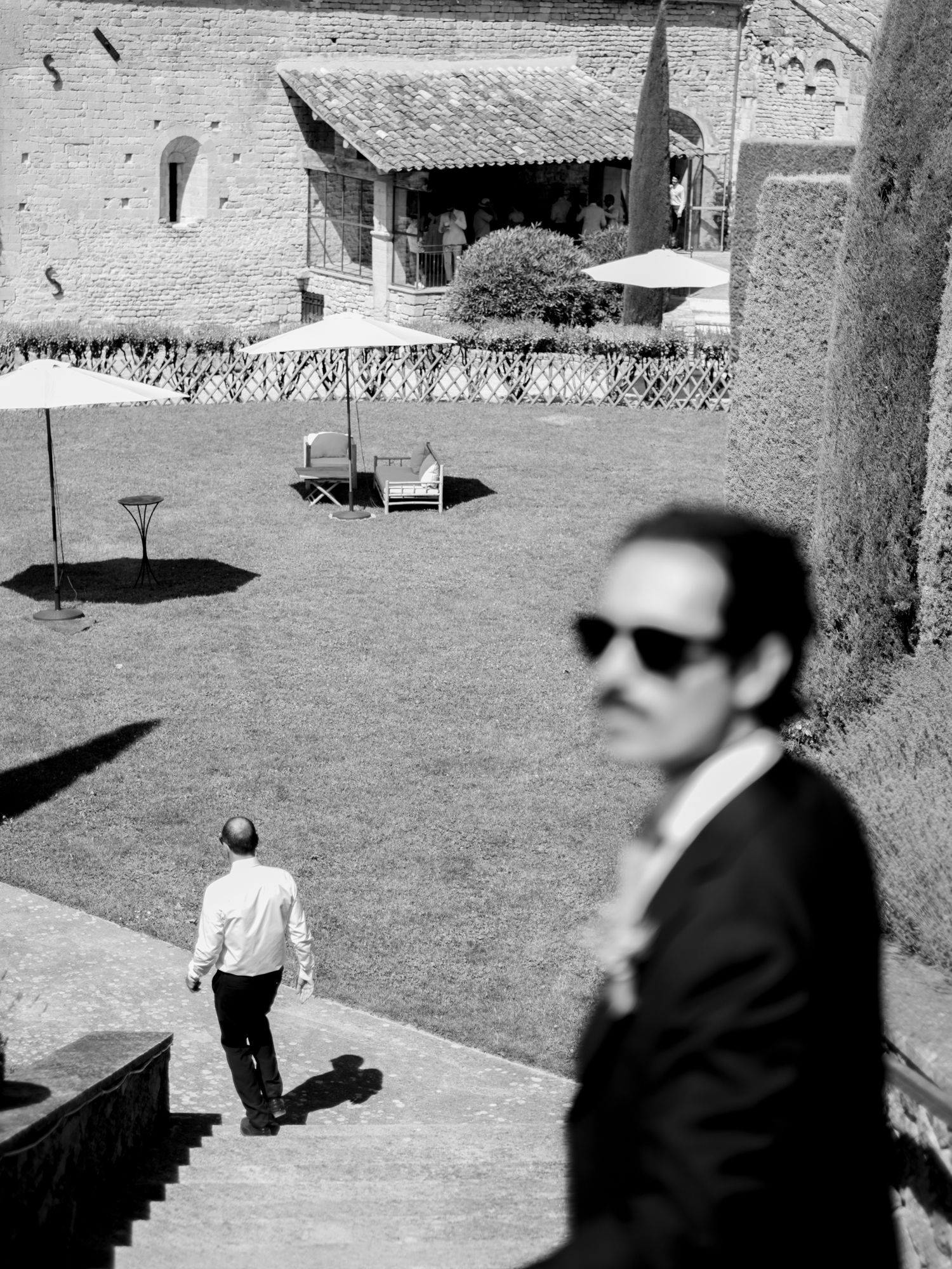 Black and white photo of two men at a historic stone villa courtyard, one in sunglasses in foreground, another walking on lawn.