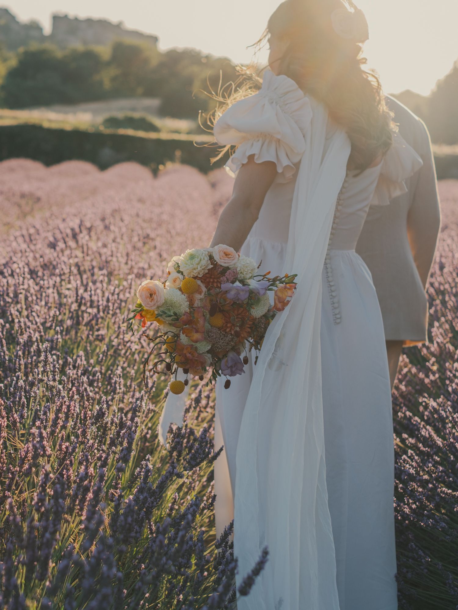 Bride in white dress with ruffled sleeves holding colorful bouquet in a lavender field at golden hour.