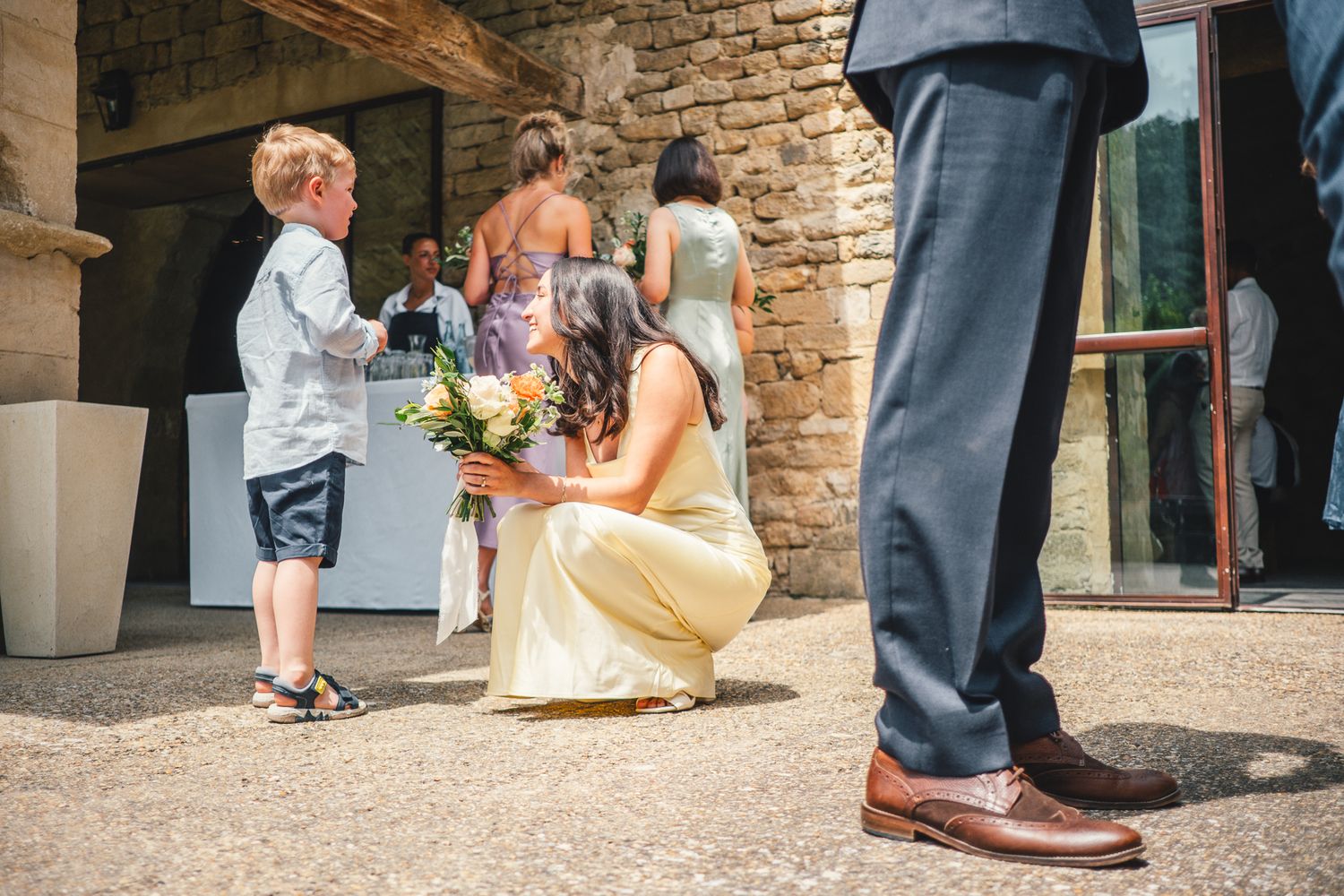 Bride crouching to greet a young boy holding flowers at an outdoor wedding venue on a sunny day.
