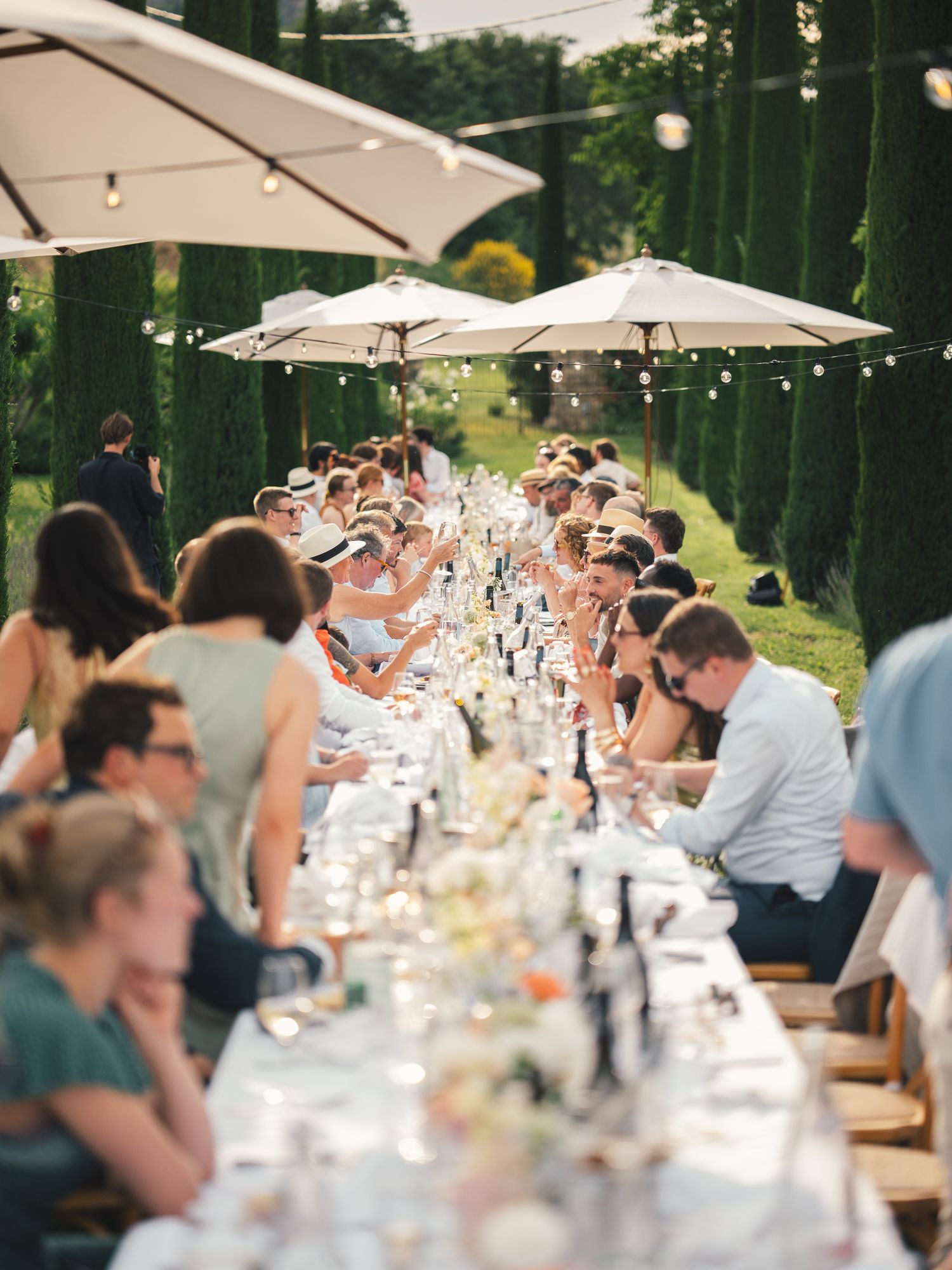 Elegant outdoor long table dinner party with guests seated under white umbrellas, string lights, and tall cypress trees.