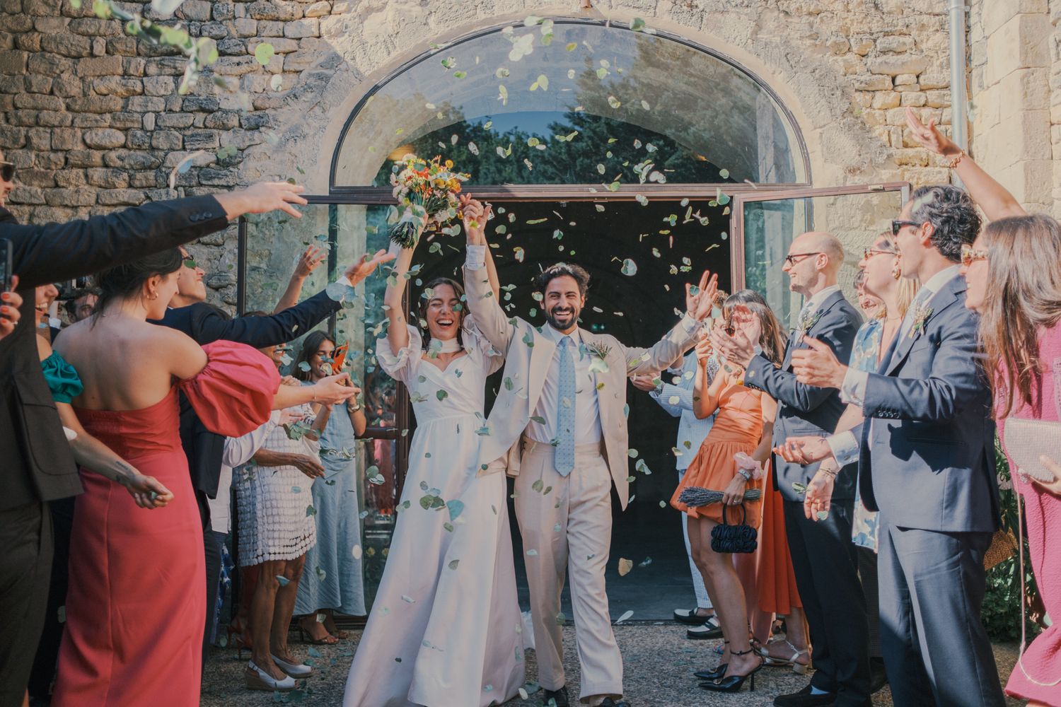 Joyful newlyweds exit stone church as guests toss colorful confetti during outdoor wedding celebration.