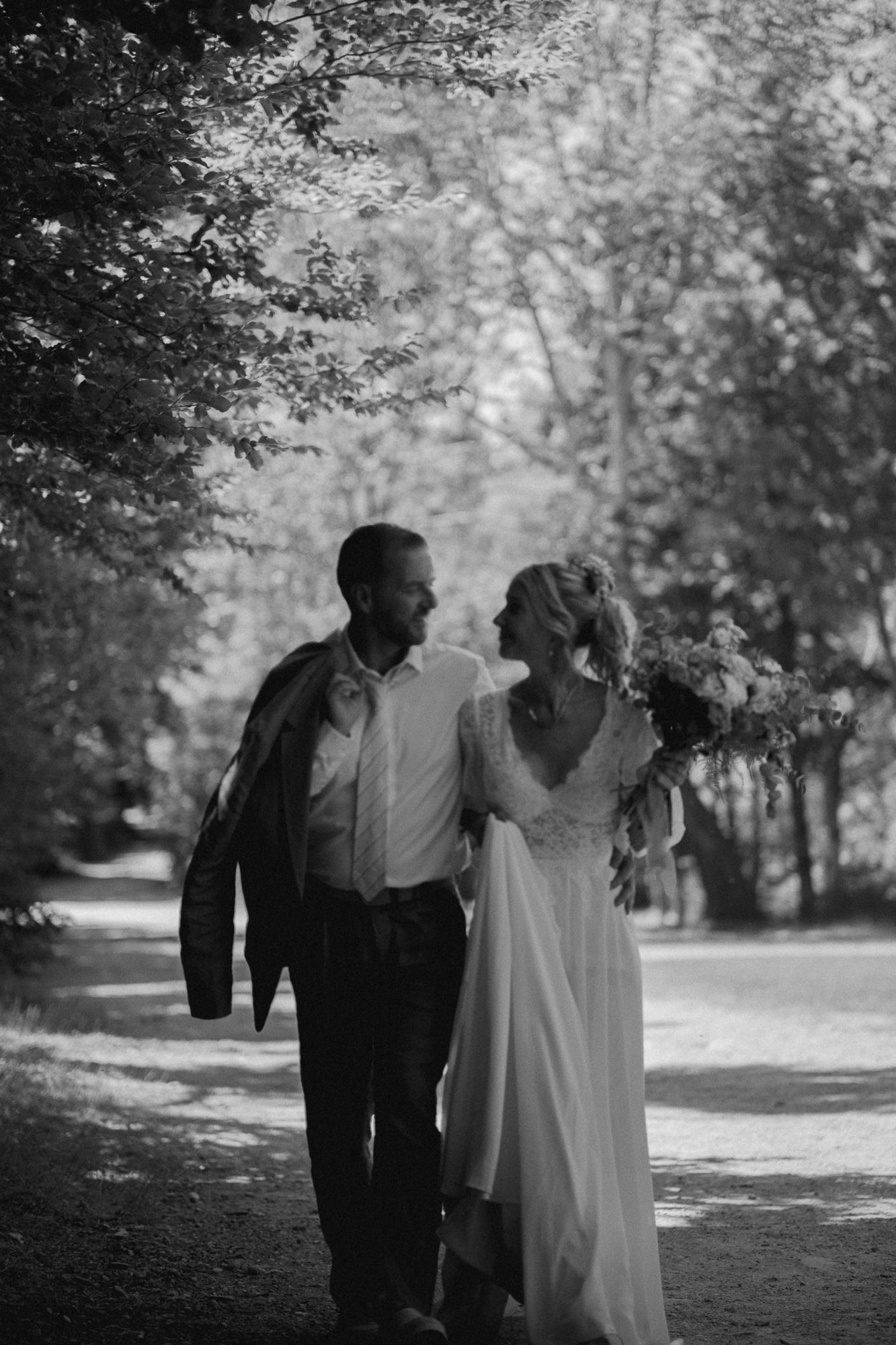 Black and white photo of a bride and groom walking together on a tree-lined path, smiling at each other.