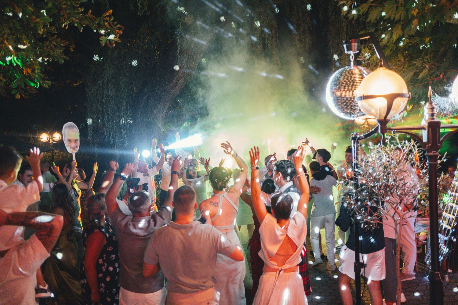 Outdoor nighttime wedding reception with guests celebrating under disco ball lights with colorful confetti and smoke effects.
