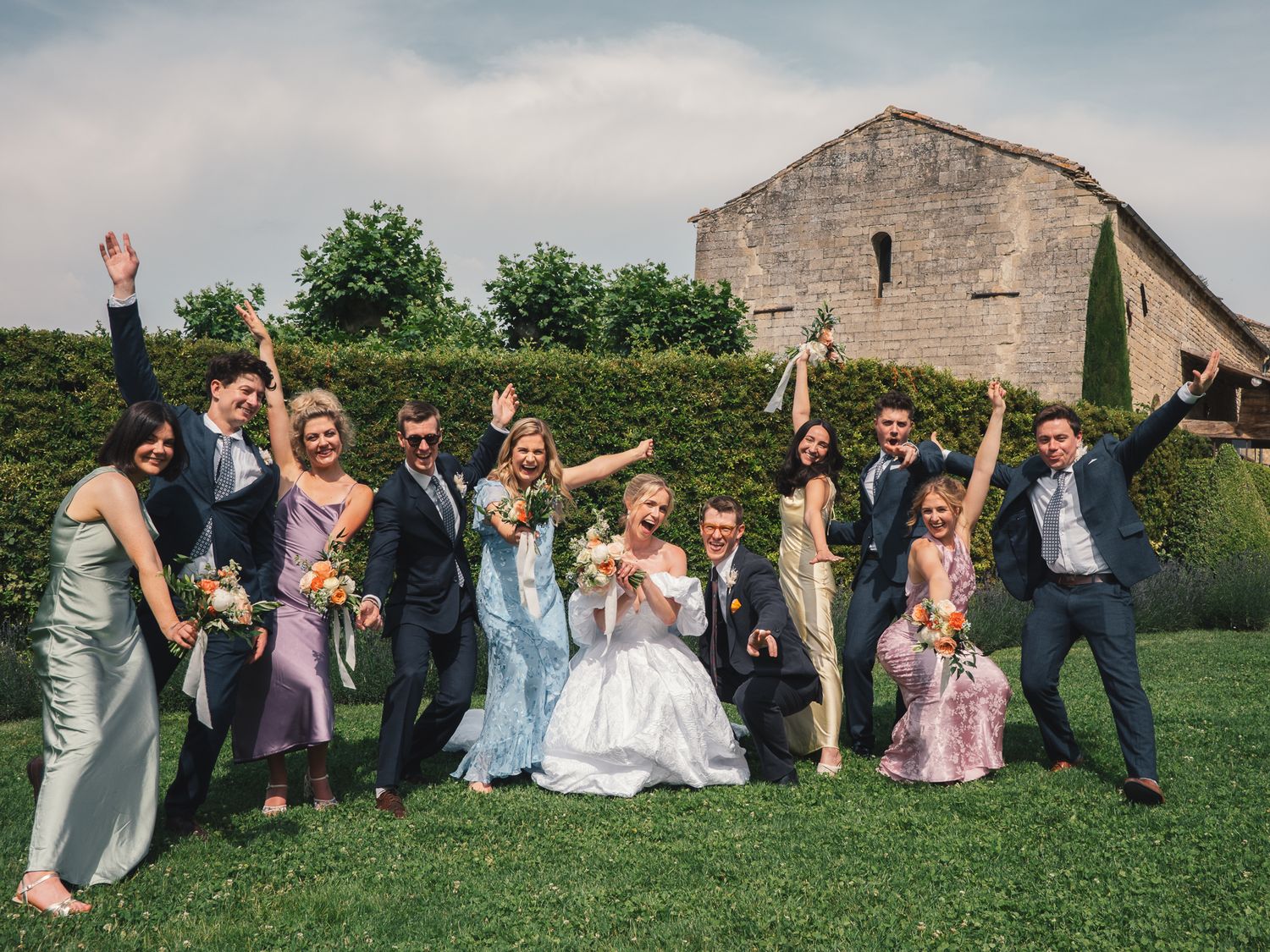 Joyful wedding party celebrating outdoors in front of a rustic stone building, with bride in white and guests in colorful formal attire.