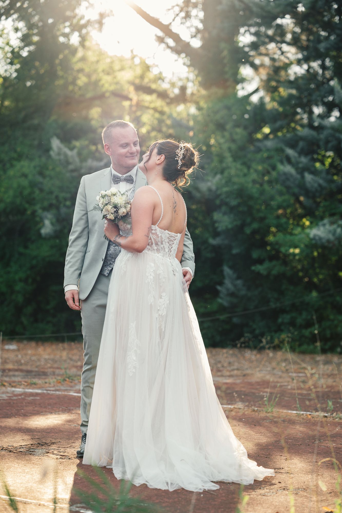 A bride and groom share a romantic moment outdoors, with golden sunlight filtering through lush green trees.