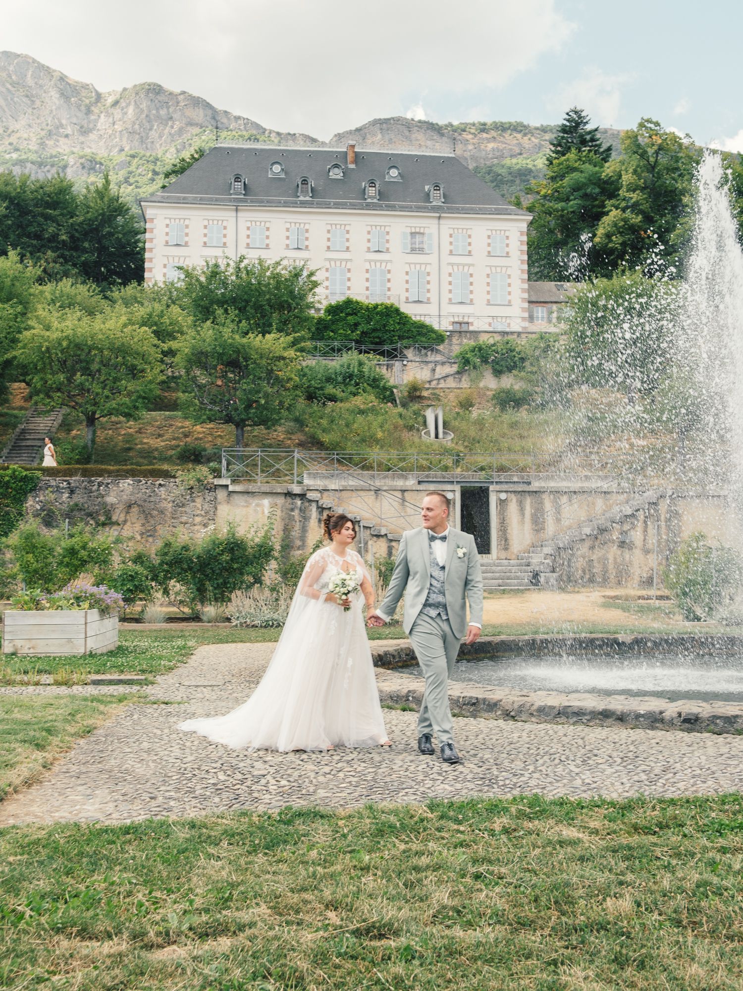 Elegant bride and groom walk hand in hand near a fountain with a grand chateau backdrop.