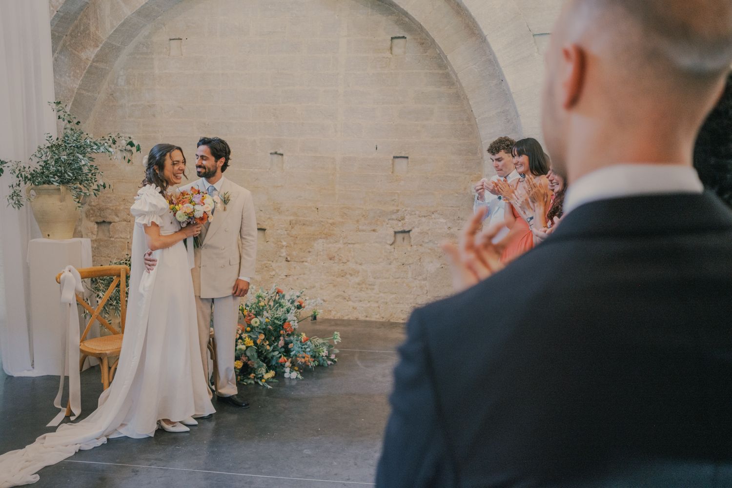 Bride and groom smiling at each other during an elegant wedding ceremony in a rustic stone venue with floral decorations.